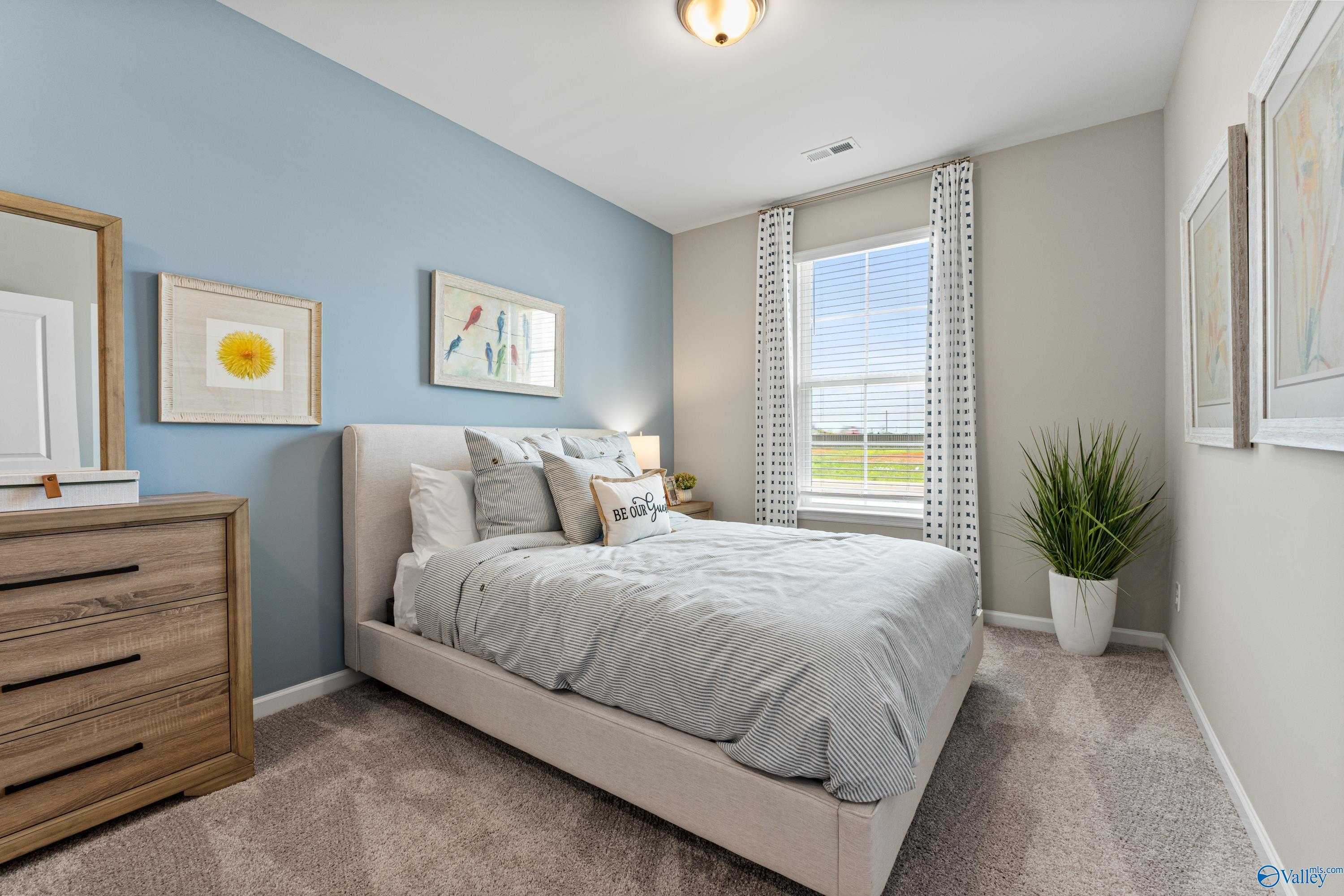 Cozy bedroom with light blue walls, upholstered queen bed, wooden dresser, and window overlooking field in Davidson Homes The Sanctuary, Athens AL