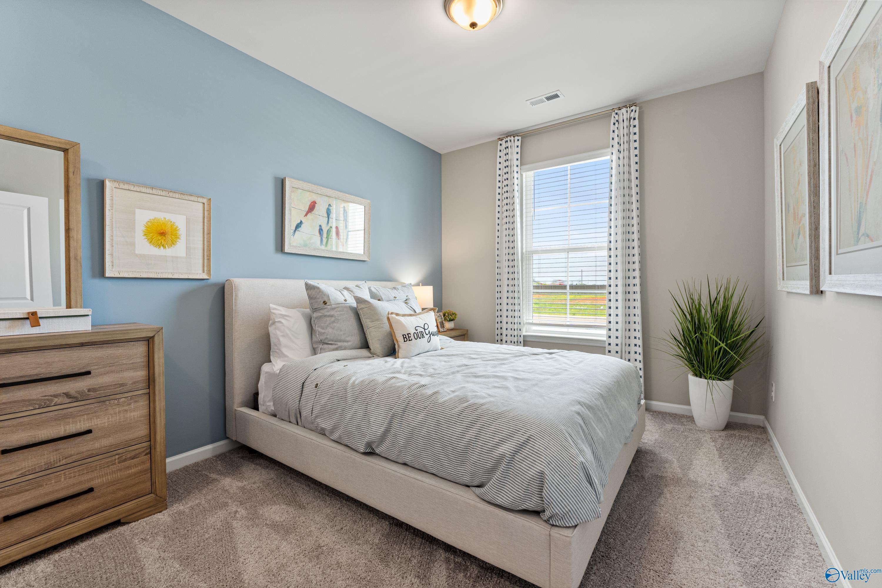 Cozy bedroom with light blue walls, upholstered queen bed, wooden dresser, and window overlooking field in Davidson Homes The Sanctuary, Athens AL