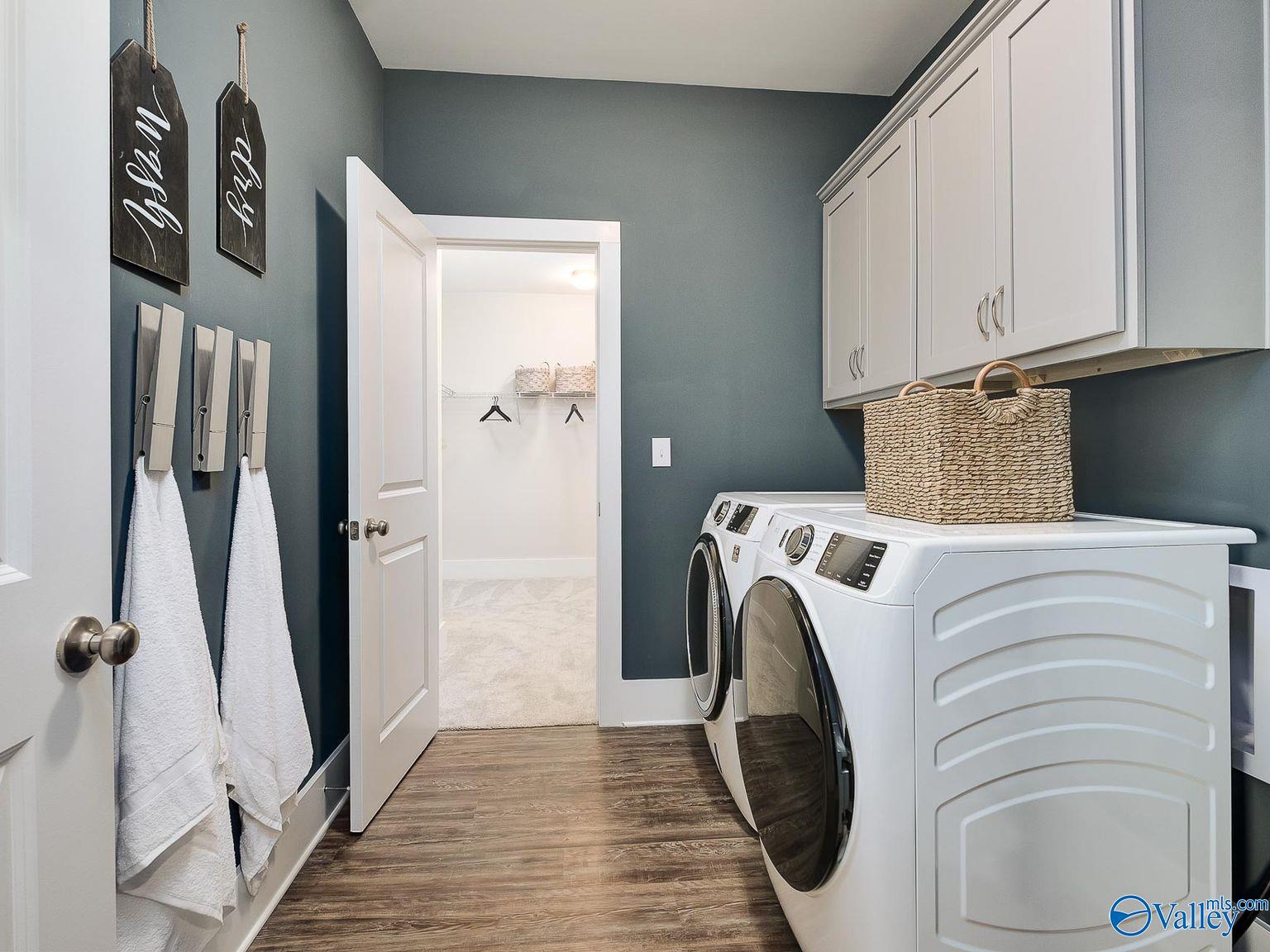 Modern laundry room with white front-load washer dryer, gray walls, white cabinets, and hanging towels in The Everett home, Toney, Alabama