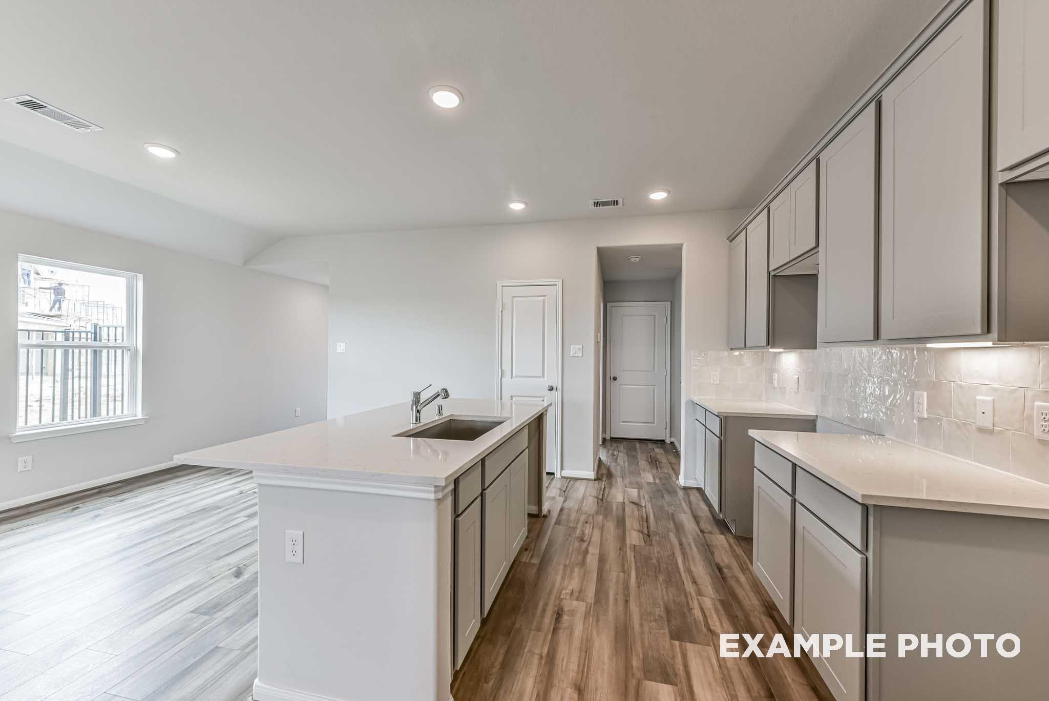 Open-concept kitchen with white shaker cabinets, large island with sink, tile backsplash, and wood-look flooring in The Costa A plan at River Ranch Meadows, Dayton, Texas