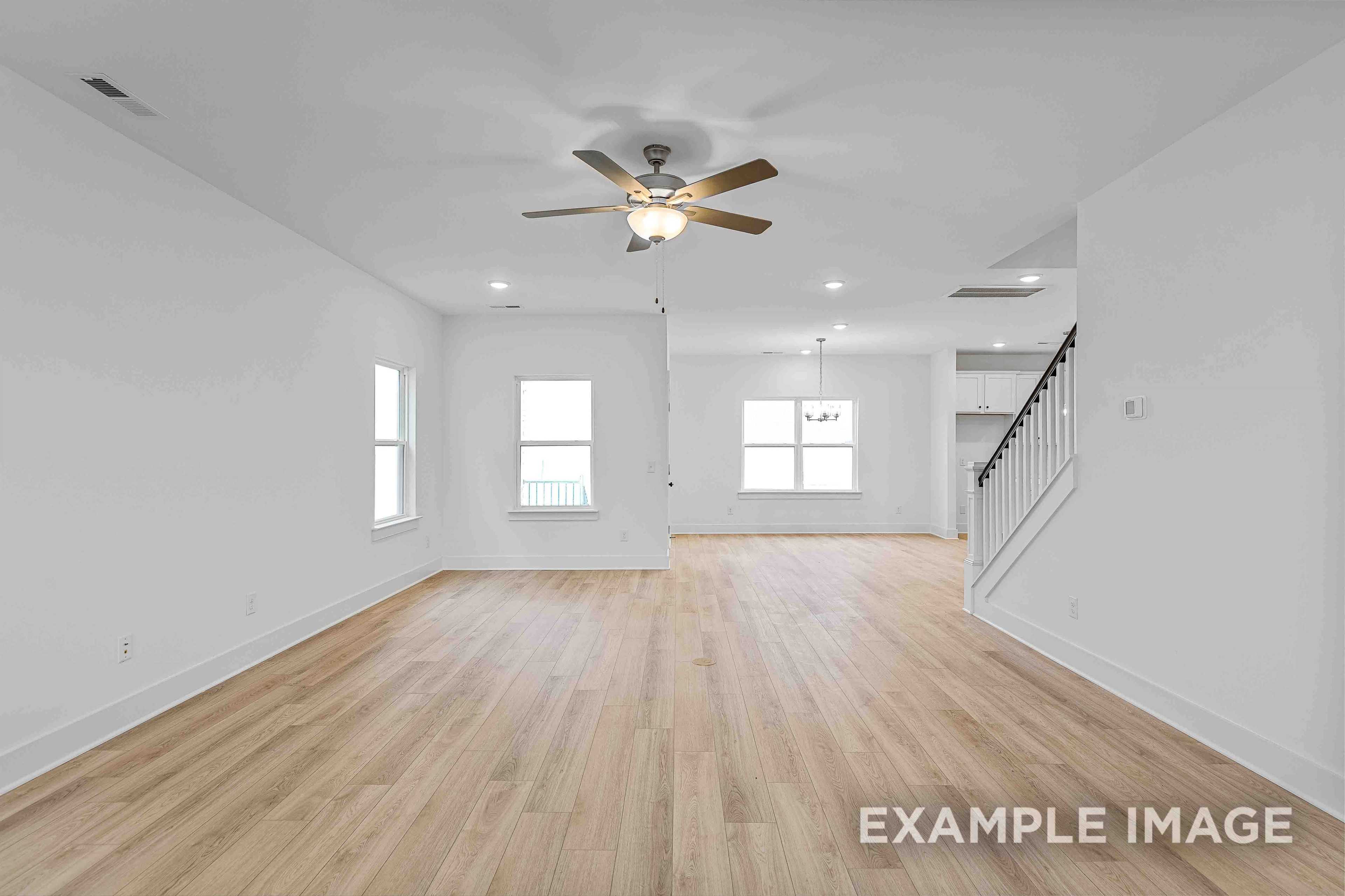 Spacious open-concept living room in The Charleston home with hardwood floors, white walls, large windows, and staircase