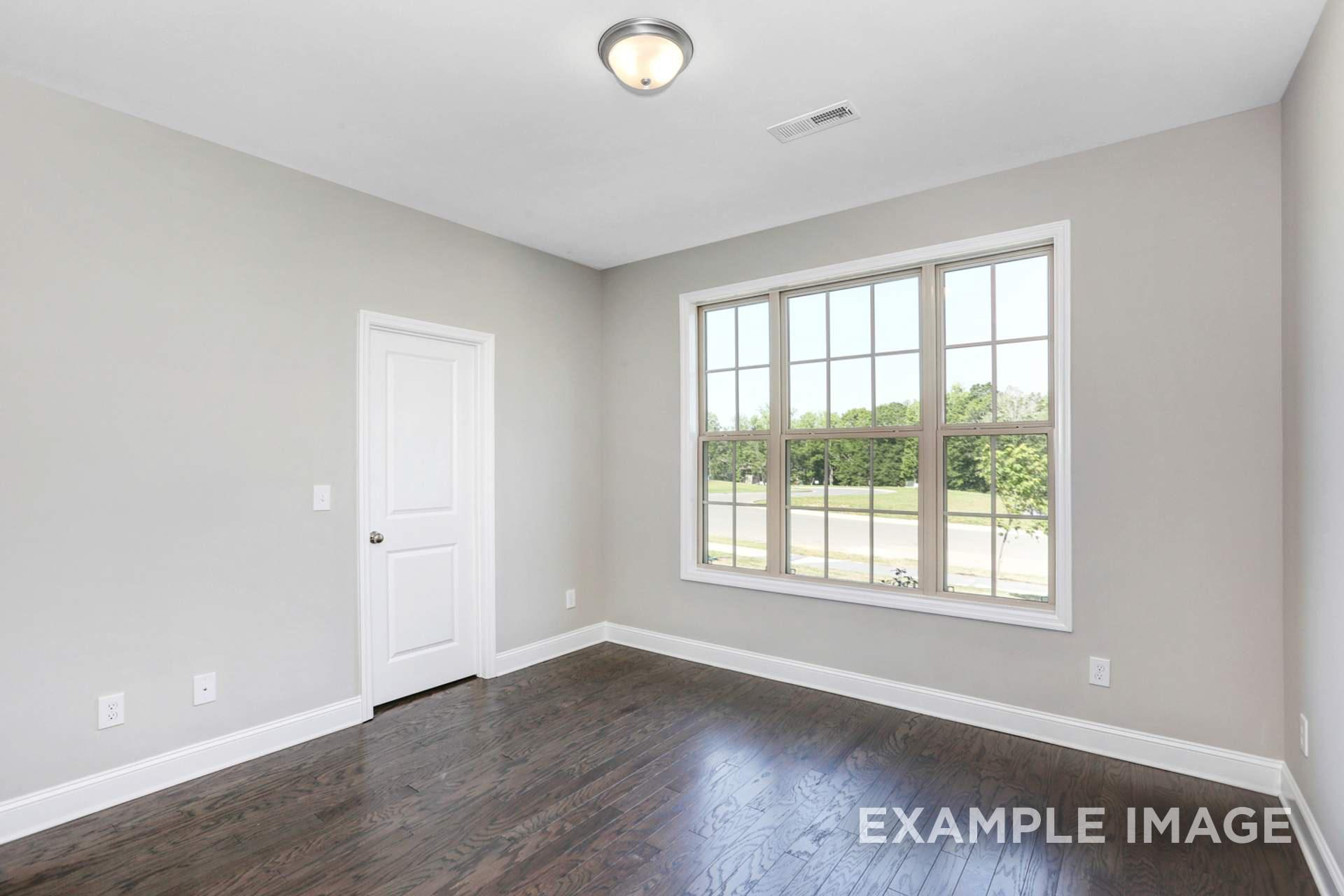 Spacious bedroom in The Montgomery B with light gray walls, dark hardwood floors, large window overlooking trees, and white door in Meridianville AL
