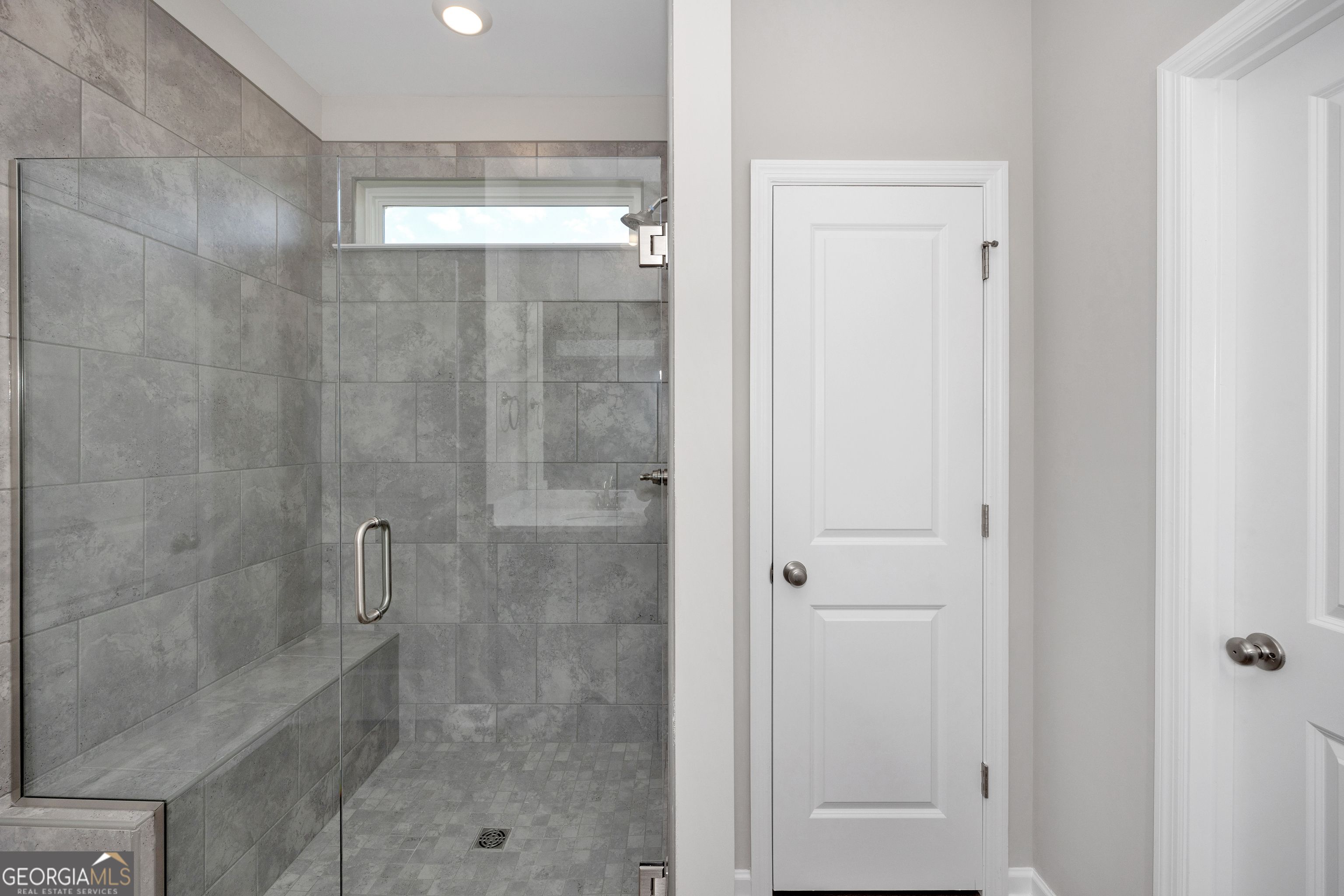 Modern master bathroom featuring frameless glass shower with gray tiles, built-in bench, and window in Evermore Homes The Luna, Perry, Georgia