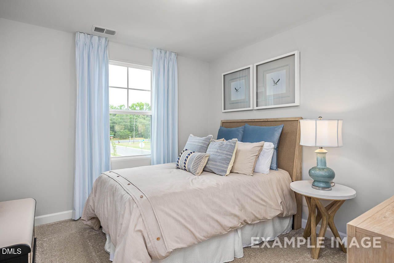Cozy secondary bedroom with queen bed, blue pillows, bedside lamp, and window view in Davidson Homes The Graham, Fuquay-Varina, NC