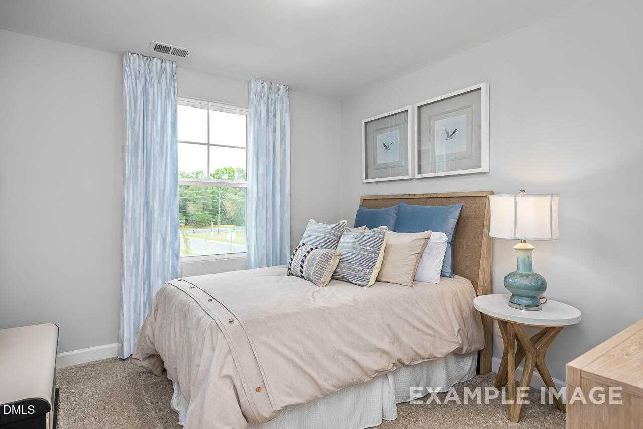 Cozy secondary bedroom featuring beige bedding, blue patterned pillows, large window with sheer curtains, and wood nightstand in Davidson Homes The Graham, Fuquay-Varina, NC