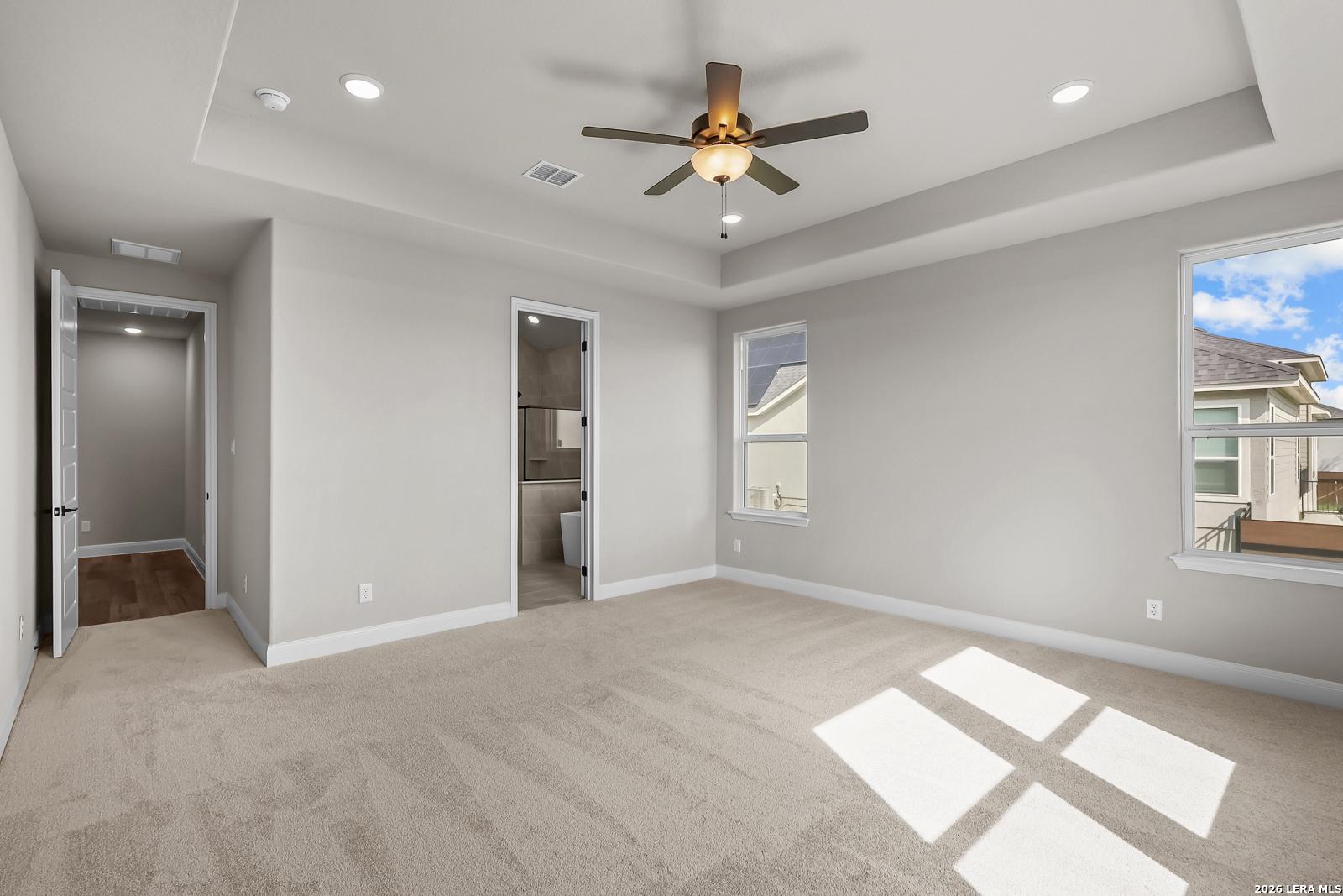 Bright primary bedroom with tray ceiling, ceiling fan, and carpet flooring in Davidson Homes The Foster E, Potranco Oaks, Castroville, TX