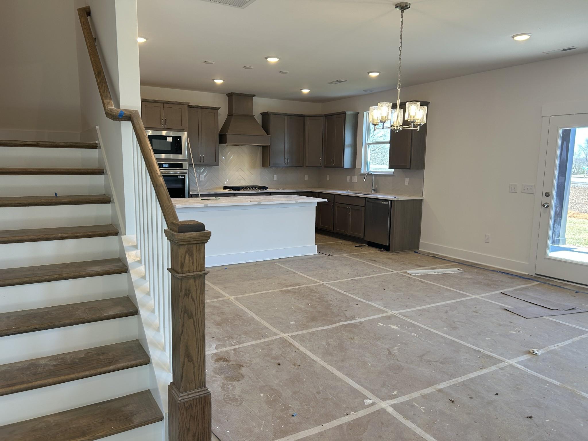 Open-concept kitchen with white shaker cabinets, stainless appliances, large island, and adjacent oak staircase in Davidson Homes The Henry C, Mt. Juliet, TN