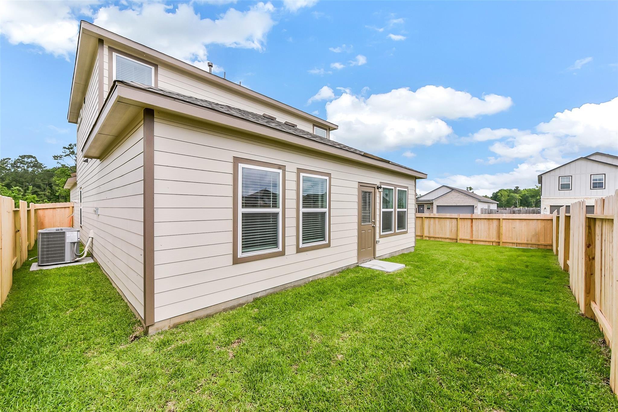 Two-story beige Sabine F home side exterior with windows, entry door, AC unit, lush green lawn, and wooden fence in Dayton Texas