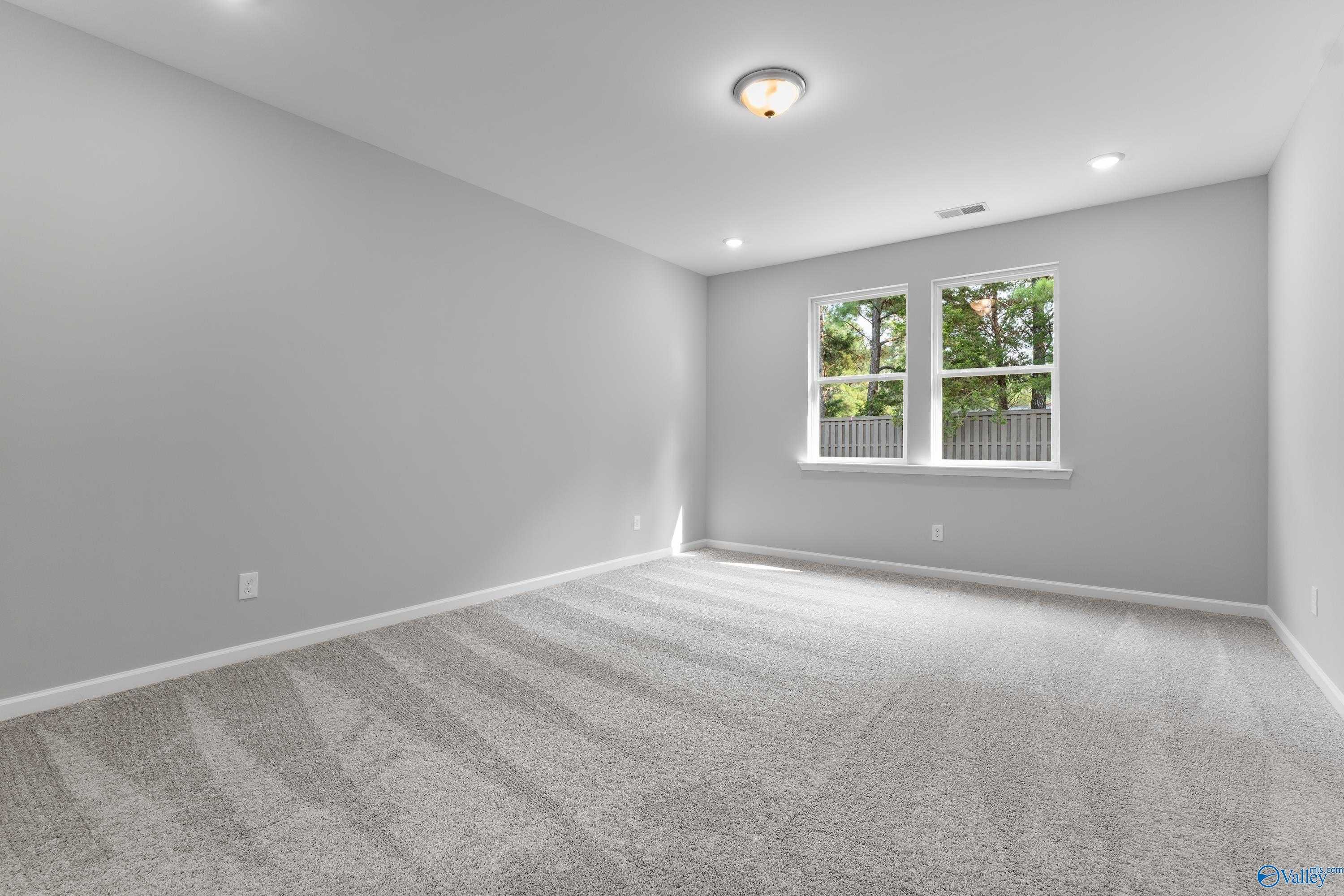 Spacious empty bedroom with light gray walls, beige carpet, and windows overlooking trees in Evermore Homes The Grace, Madison, Alabama