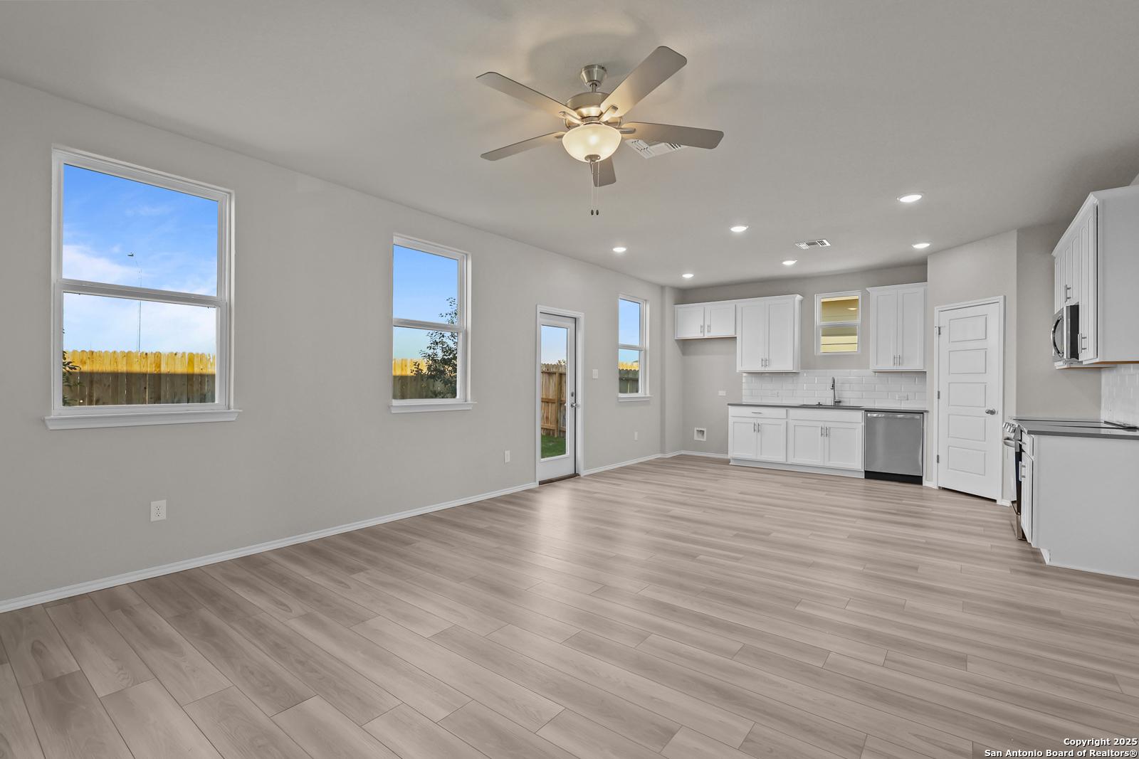 Bright open-concept kitchen-living area with white cabinets, wood floors, ceiling fan, and large windows in Davidson Homes The Trinity D, San Antonio, Texas
