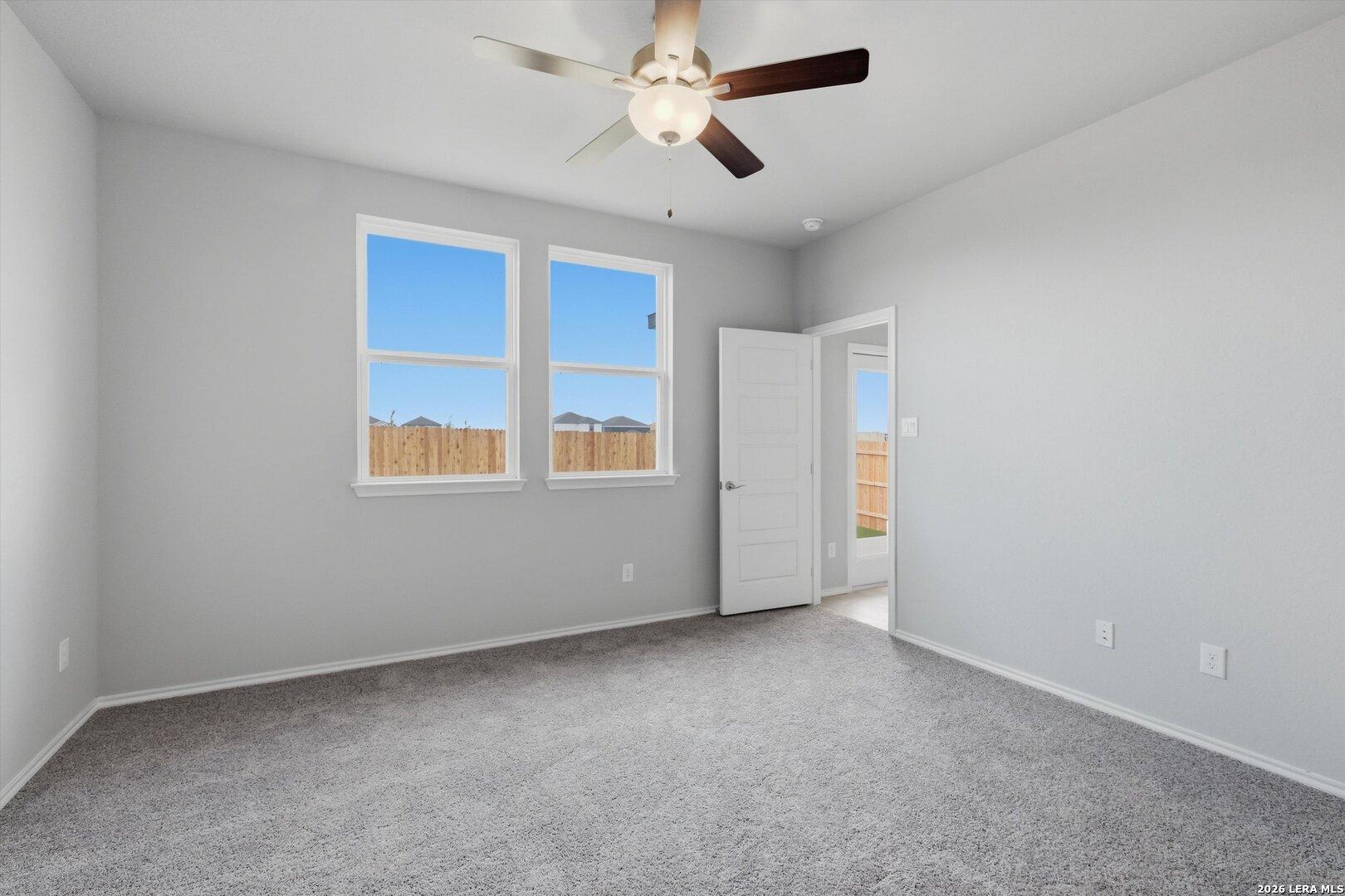 Spacious bedroom with gray walls, carpet floor, ceiling fan, and large windows overlooking fenced backyard in Davidson Homes The Frio B, San Antonio