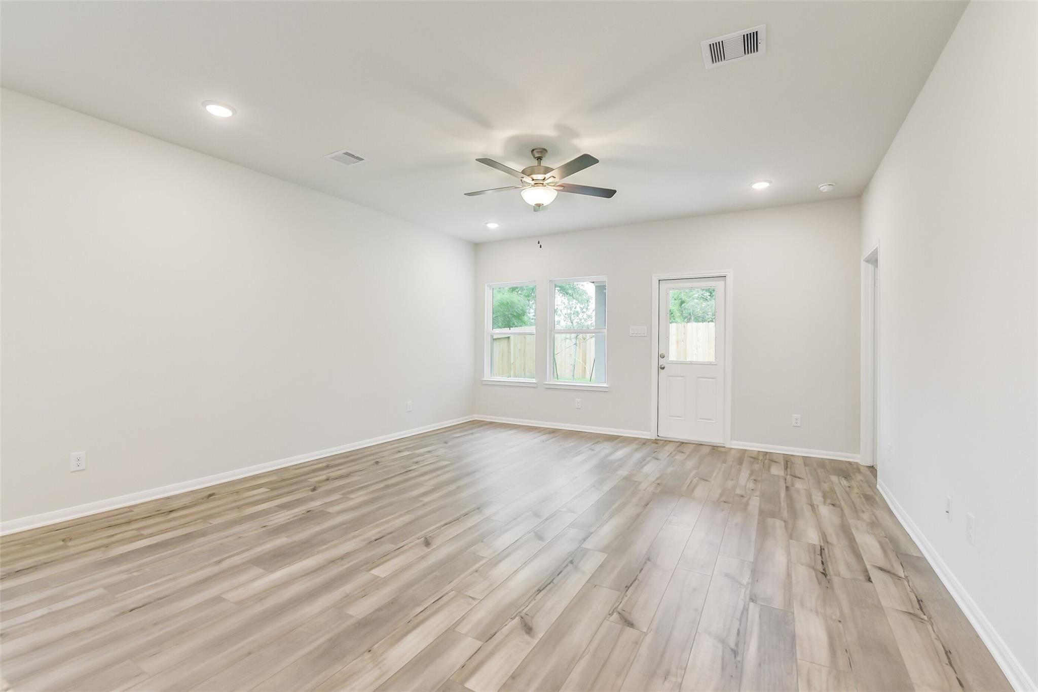 Spacious empty living room with light wood floors, ceiling fan, large windows, and white walls in Davidson Homes The Brazos F, Conroe, Texas