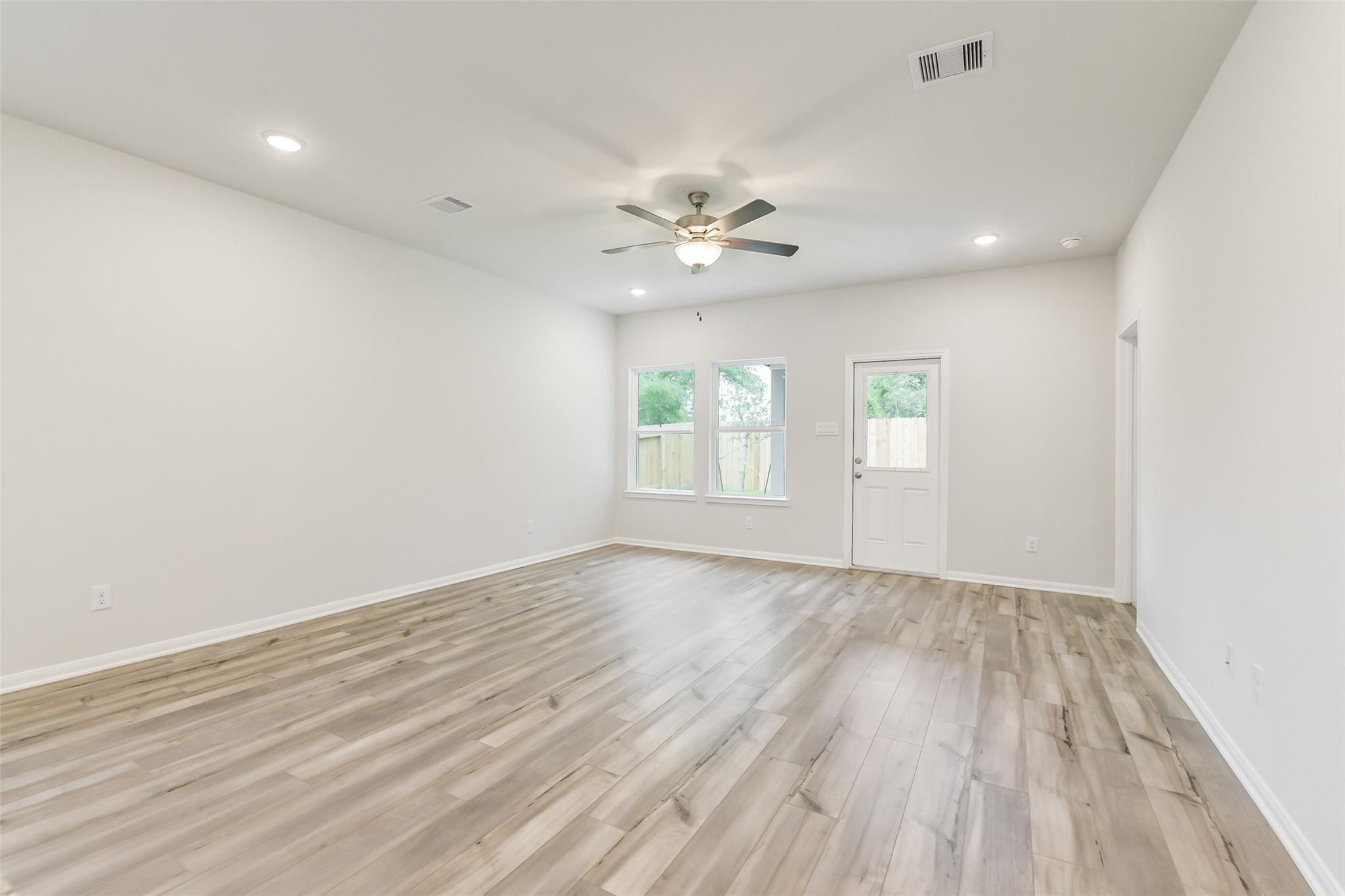 Spacious living room with light hardwood floors, ceiling fan, large windows, and white walls in 5-bedroom Davidson Homes The Brazos F, Conroe, Texas