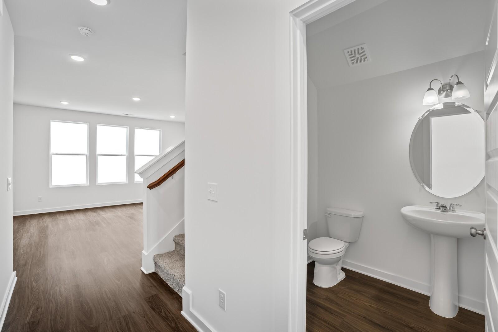Elegant hallway with hardwood floors, white staircase, and adjacent powder room featuring pedestal sink, round mirror in The Logan C, White House, TN