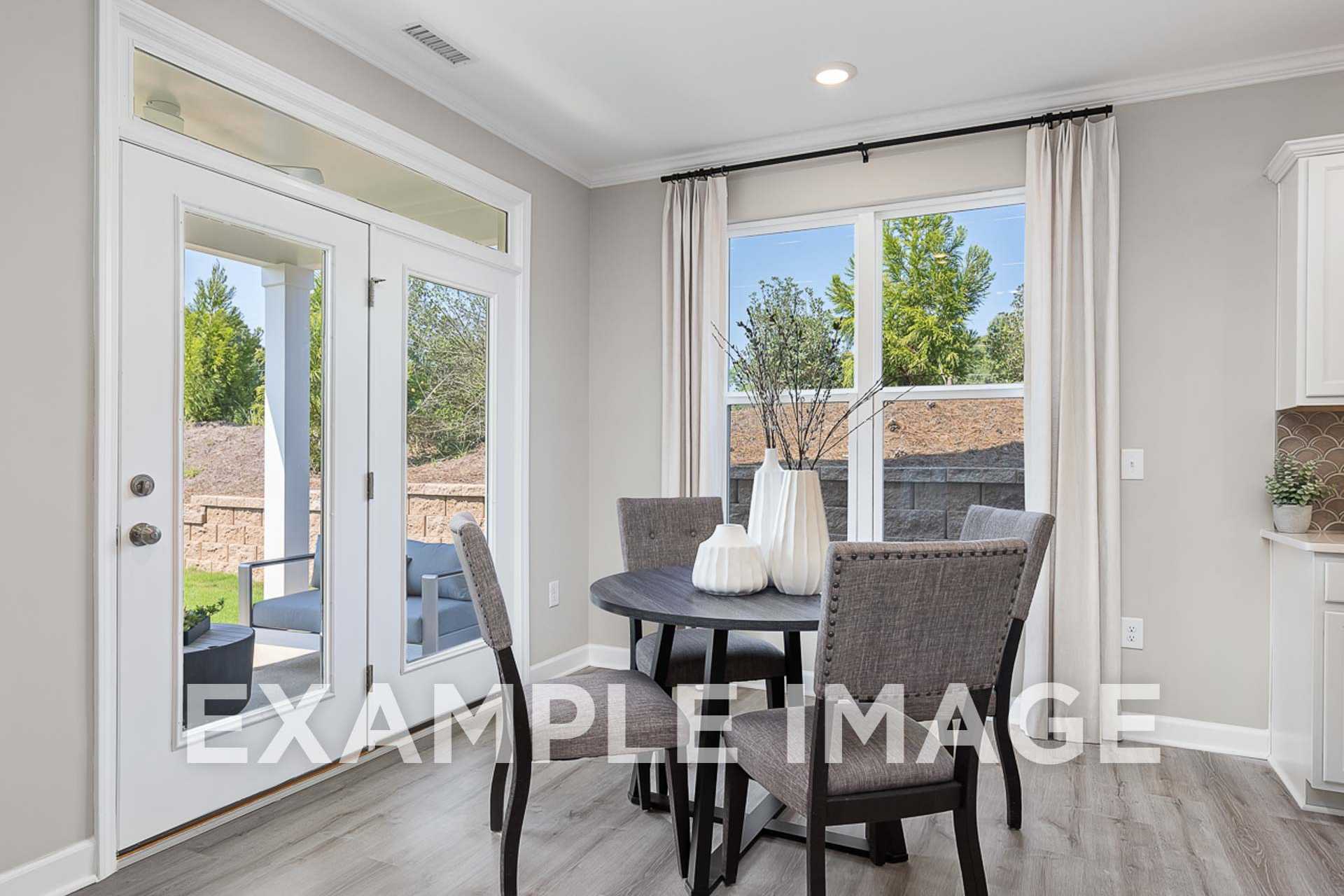 Spacious dining room in The Ash E with round wood table, upholstered chairs, French doors to tree-lined patio, and gray walls
