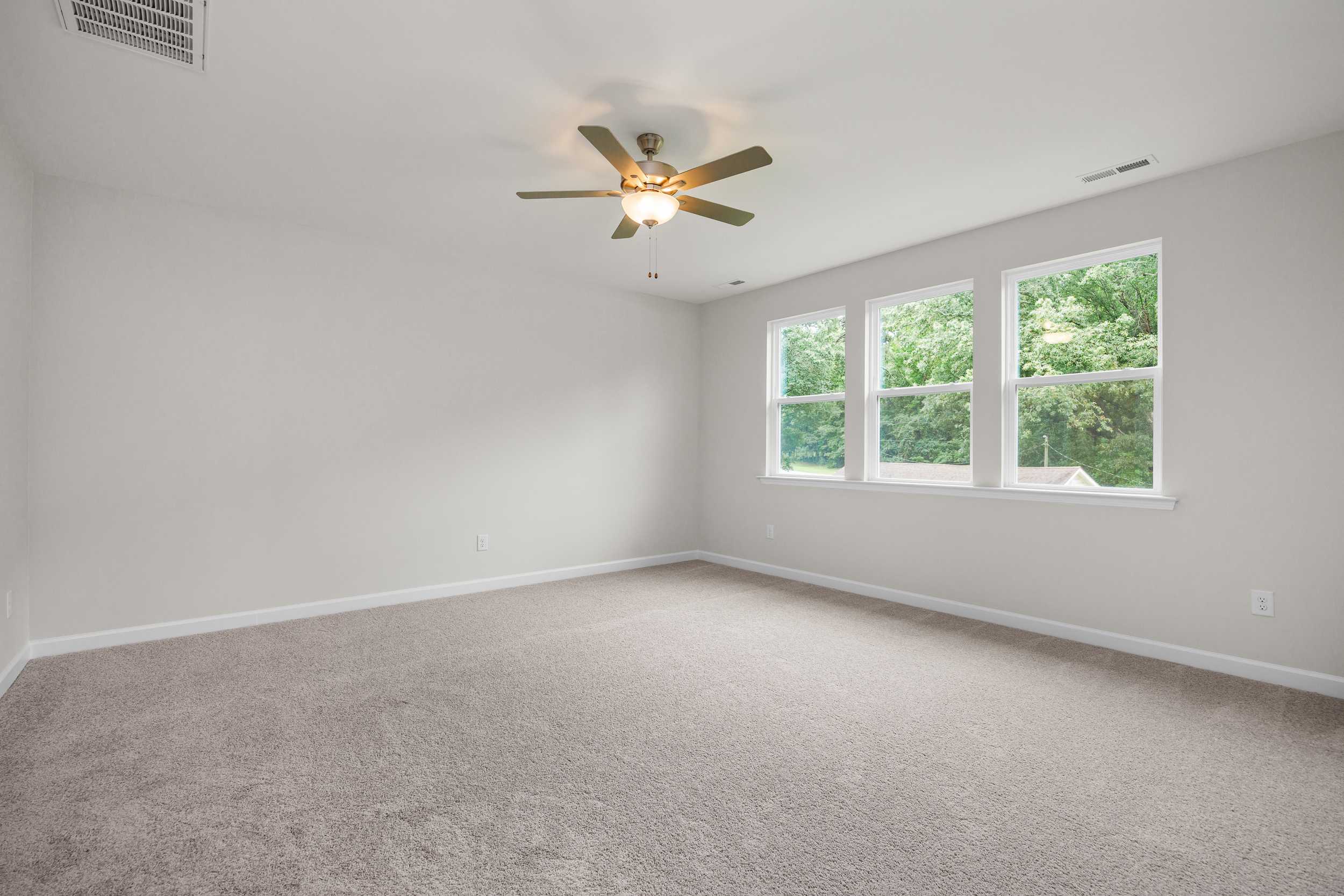 Spacious master bedroom in The Aspen with beige carpet, ceiling fan, and large triple windows overlooking trees