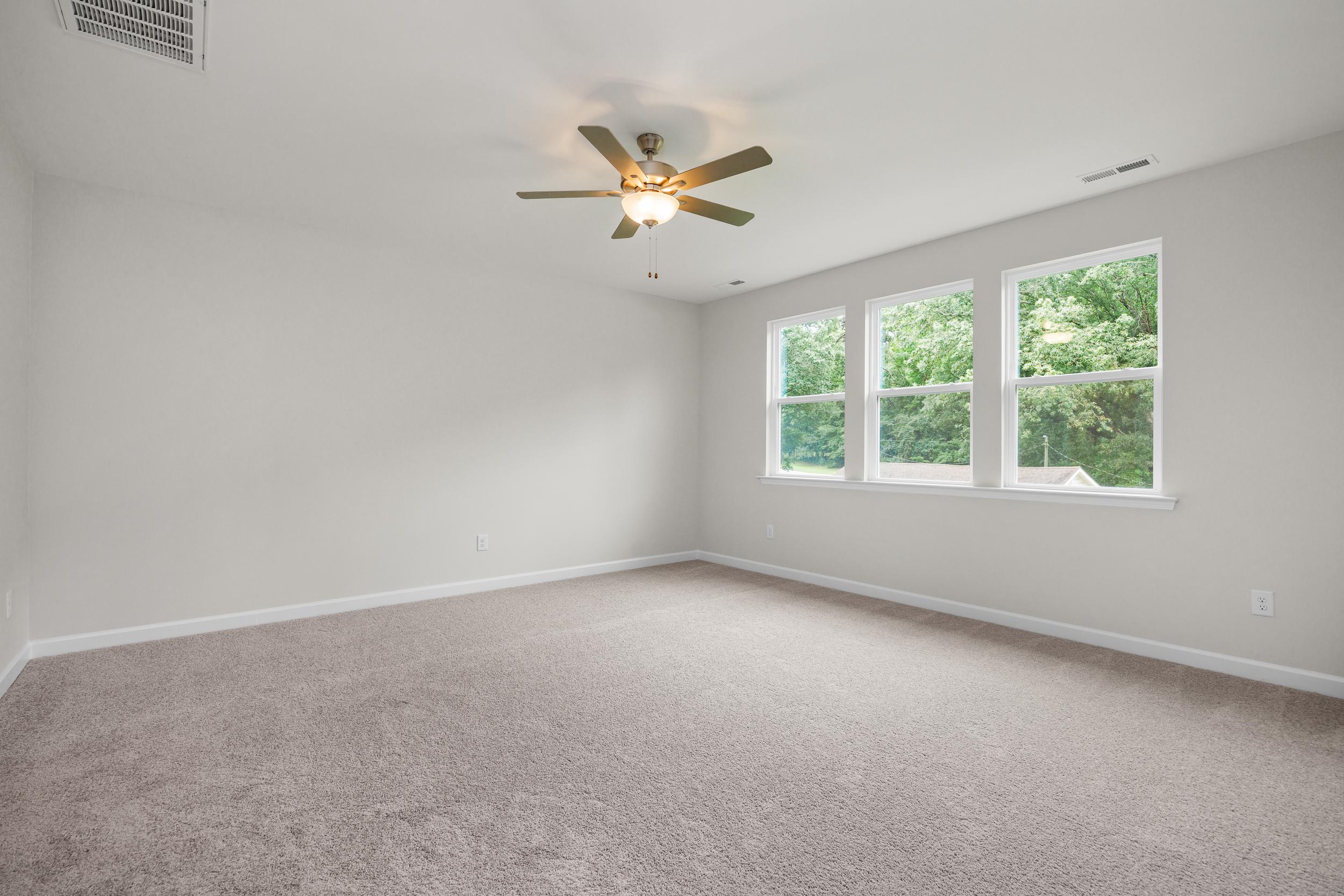 Spacious master bedroom in The Aspen with beige carpet, ceiling fan, and large triple windows overlooking trees