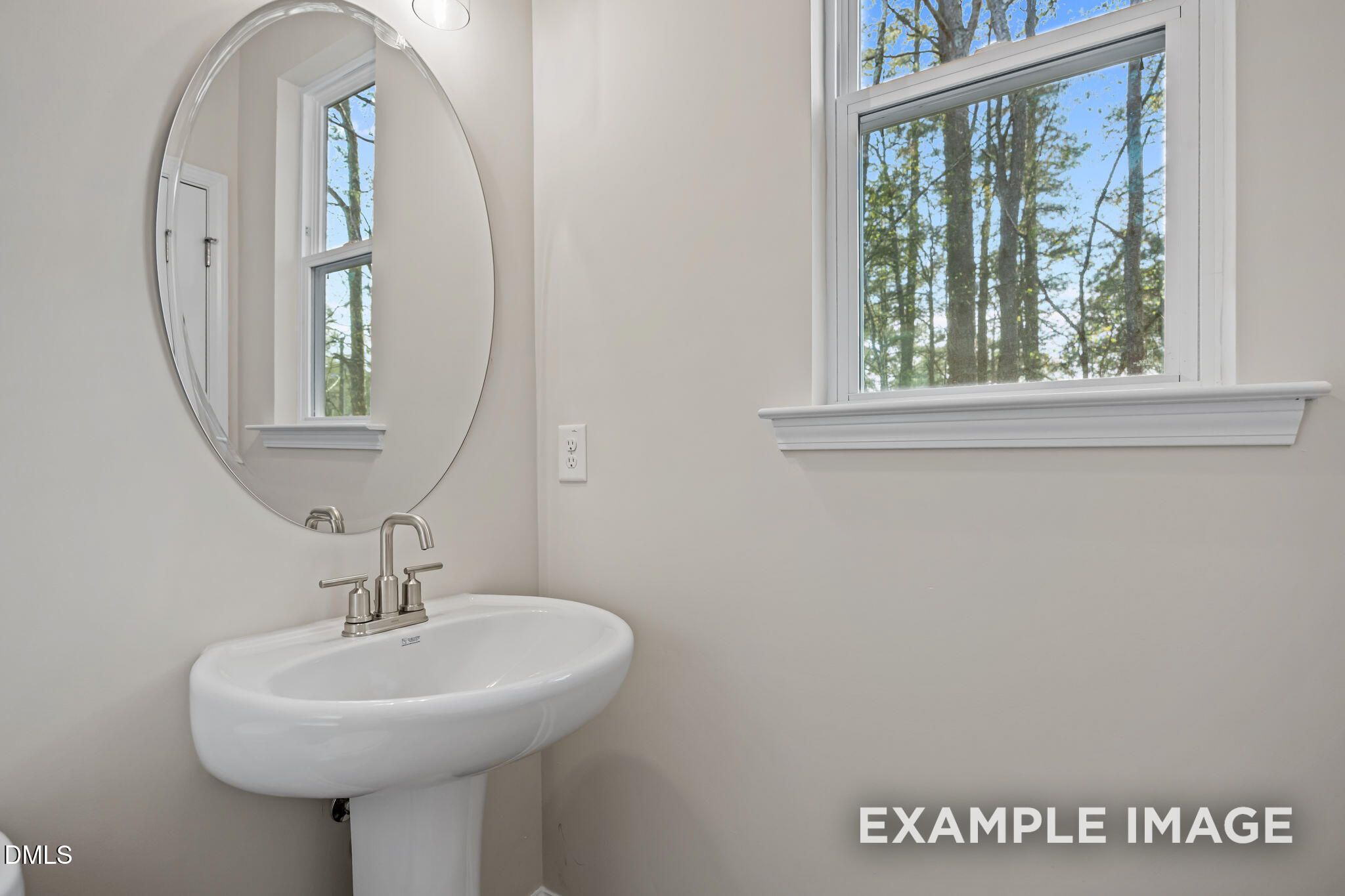Modern guest bathroom with white pedestal sink, chrome faucet, round mirror, and window view of trees in Davidson Homes The Avery, Knightdale, NC