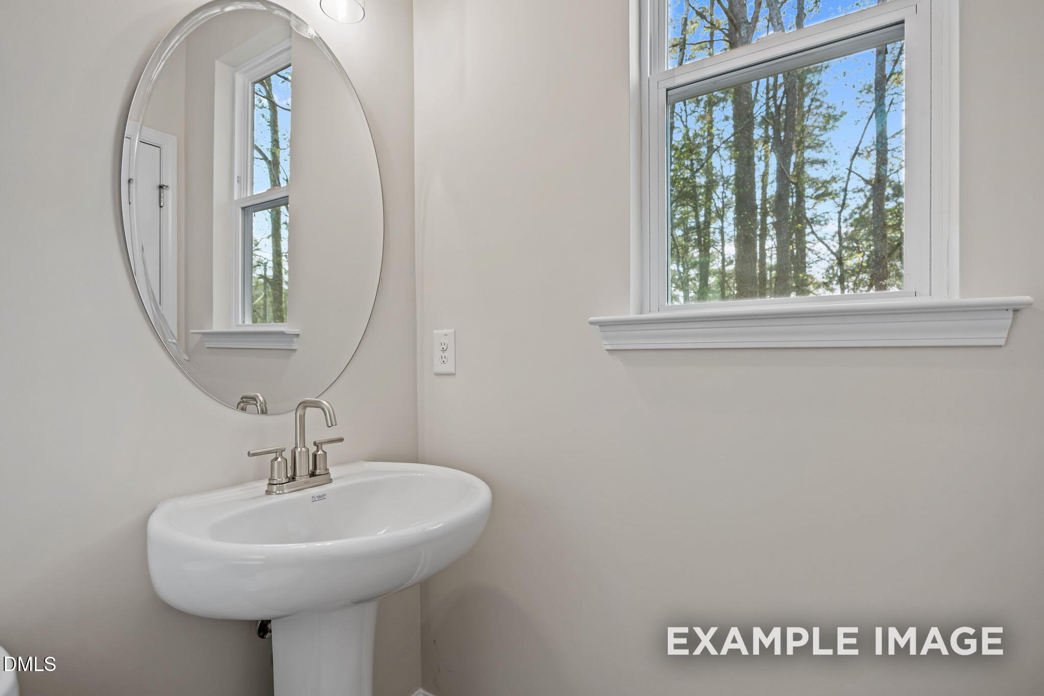Modern guest bathroom with white pedestal sink, chrome faucet, round mirror, and window view of trees in Davidson Homes The Avery, Knightdale, NC