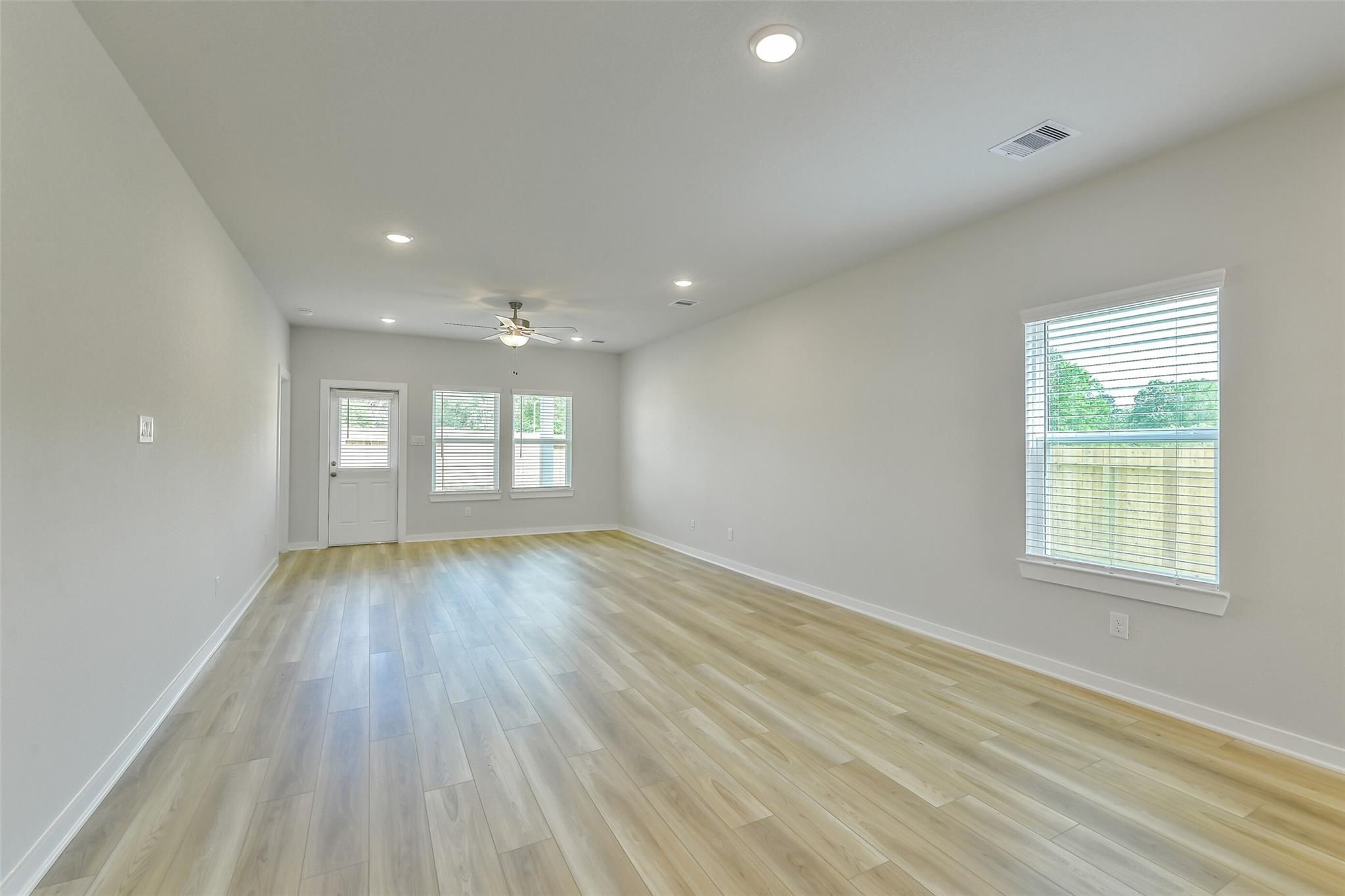 Spacious empty living room with light laminate floors, ceiling fan, and large windows overlooking backyard in The San Marcos E, Conroe, Texas