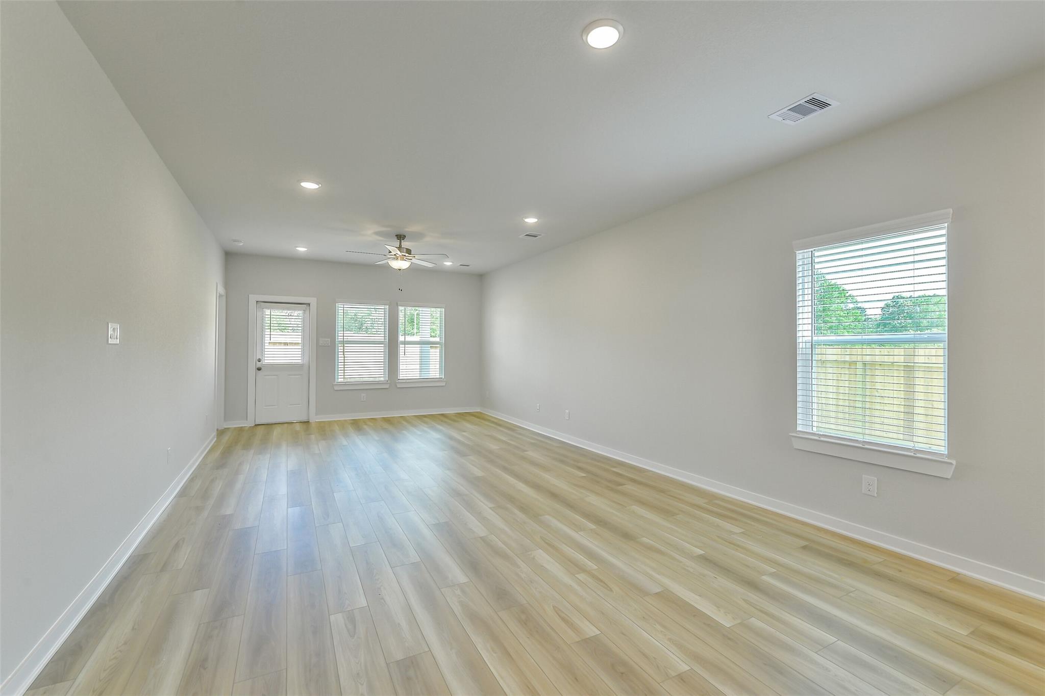 Spacious empty living room with light laminate floors, ceiling fan, and large windows overlooking backyard in The San Marcos E, Conroe, Texas