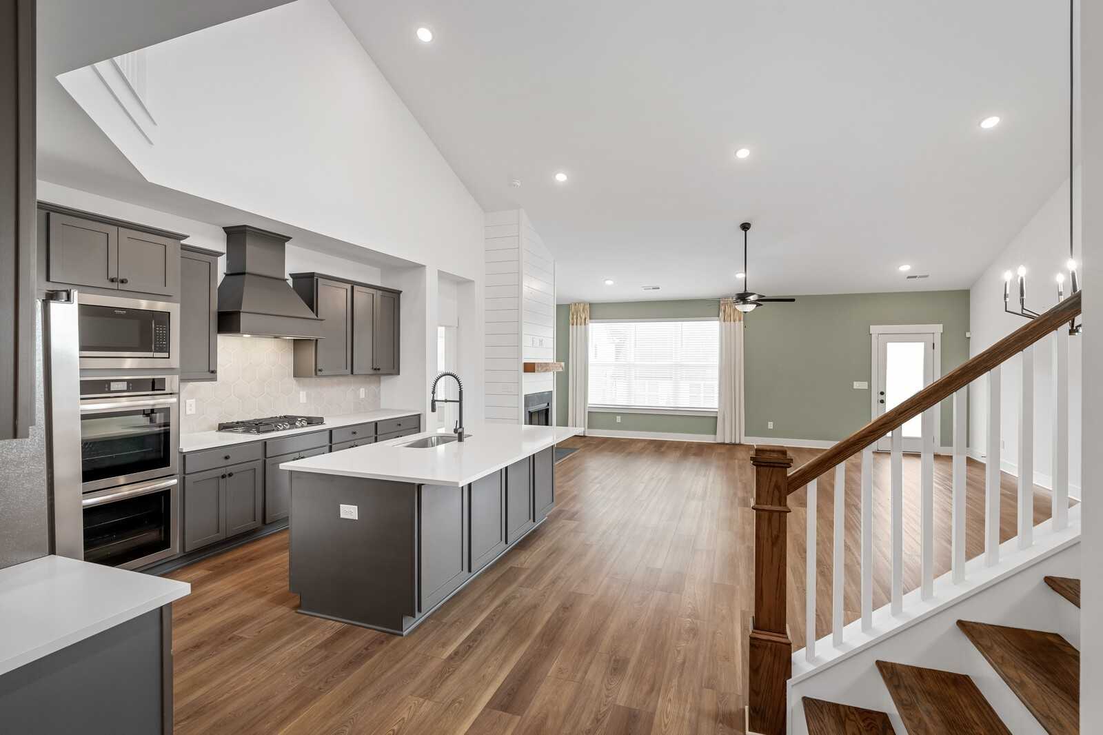 Open-concept kitchen with gray cabinets, white island, stainless ovens, and open family room with fireplace and staircase in Davidson Homes Ridgeport, Gallatin, TN