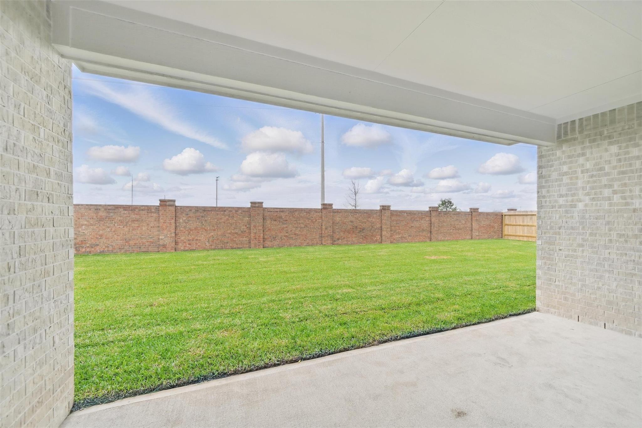 Covered back patio with concrete floor overlooking lush green lawn and brick fence under partly cloudy sky in Davidson Homes The Victoria C, Lago Mar, Texas City