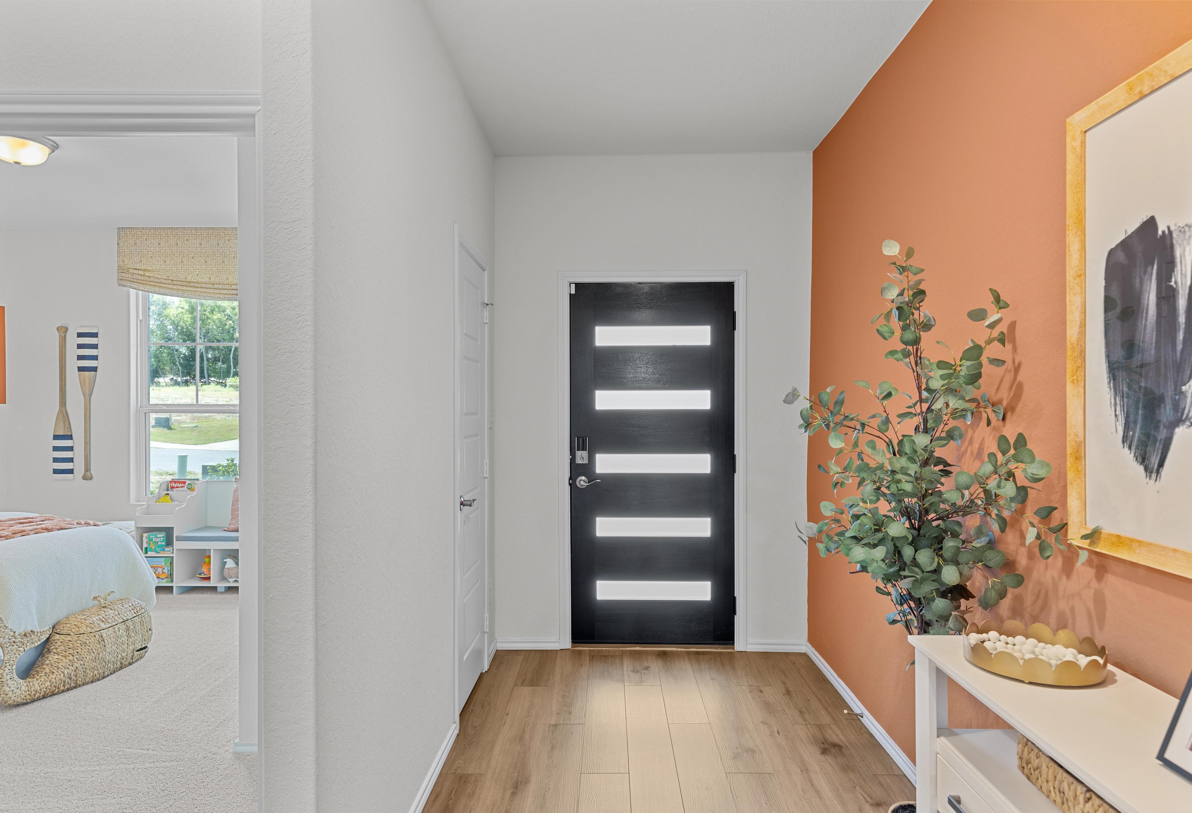 Spacious entry hallway in The Daphne home design with modern black door, orange accent wall, eucalyptus plant, and light wood floors