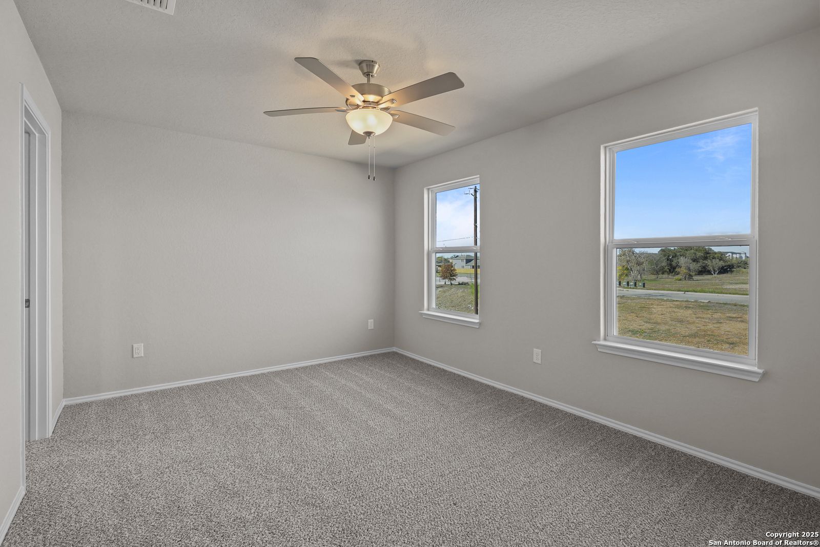 Bright bedroom with gray walls, carpet floor, ceiling fan, and large windows showing scenic fields in Davidson Homes Trinity D, San Antonio