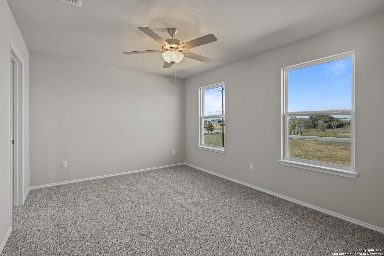 Bright bedroom with gray walls, carpet floor, ceiling fan, and large windows showing scenic fields in Davidson Homes Trinity D, San Antonio