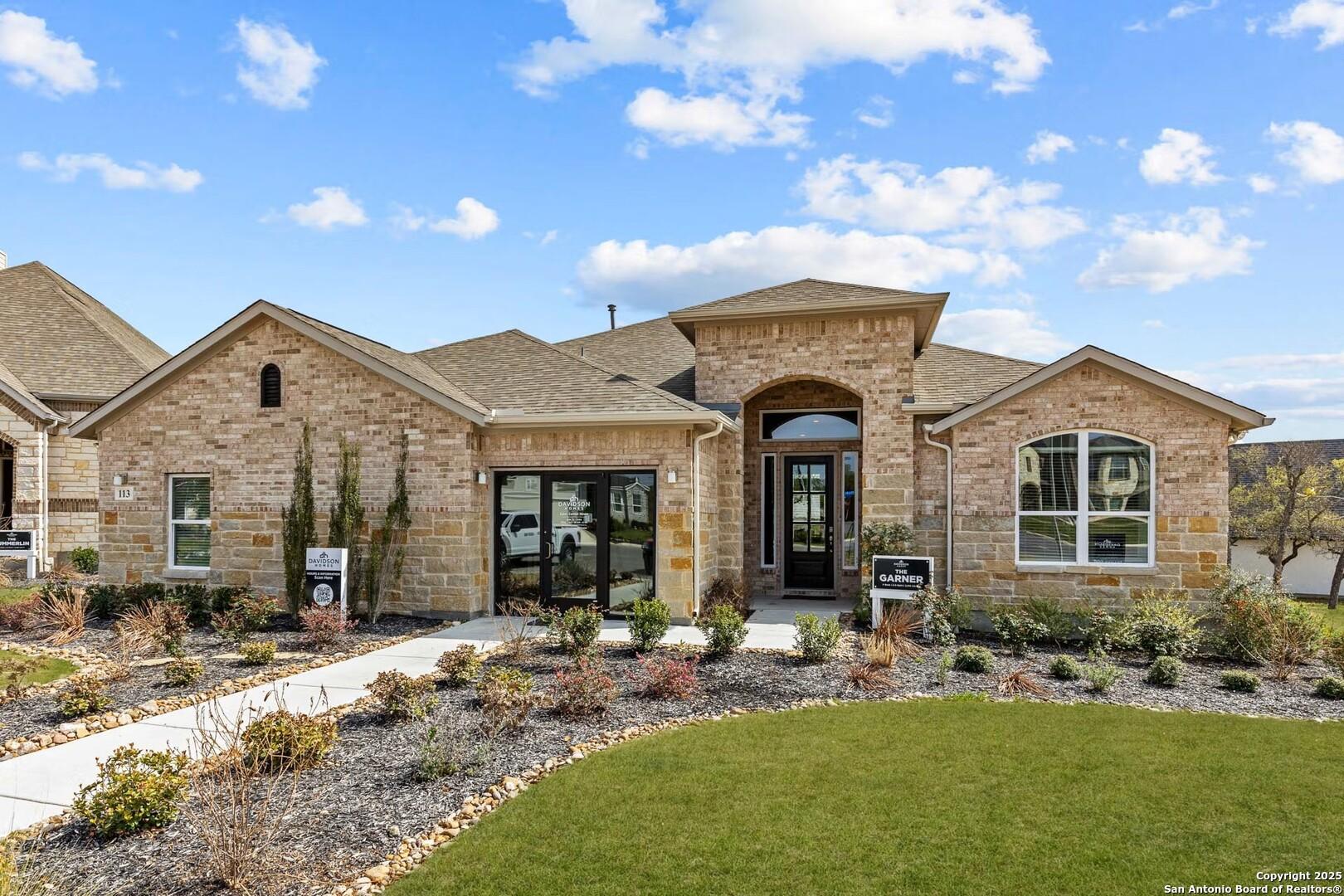 Tan brick single-story home with arched entry, 3-car garage, and landscaped yard in The Reserve at Potranco Oaks, Castroville, Texas