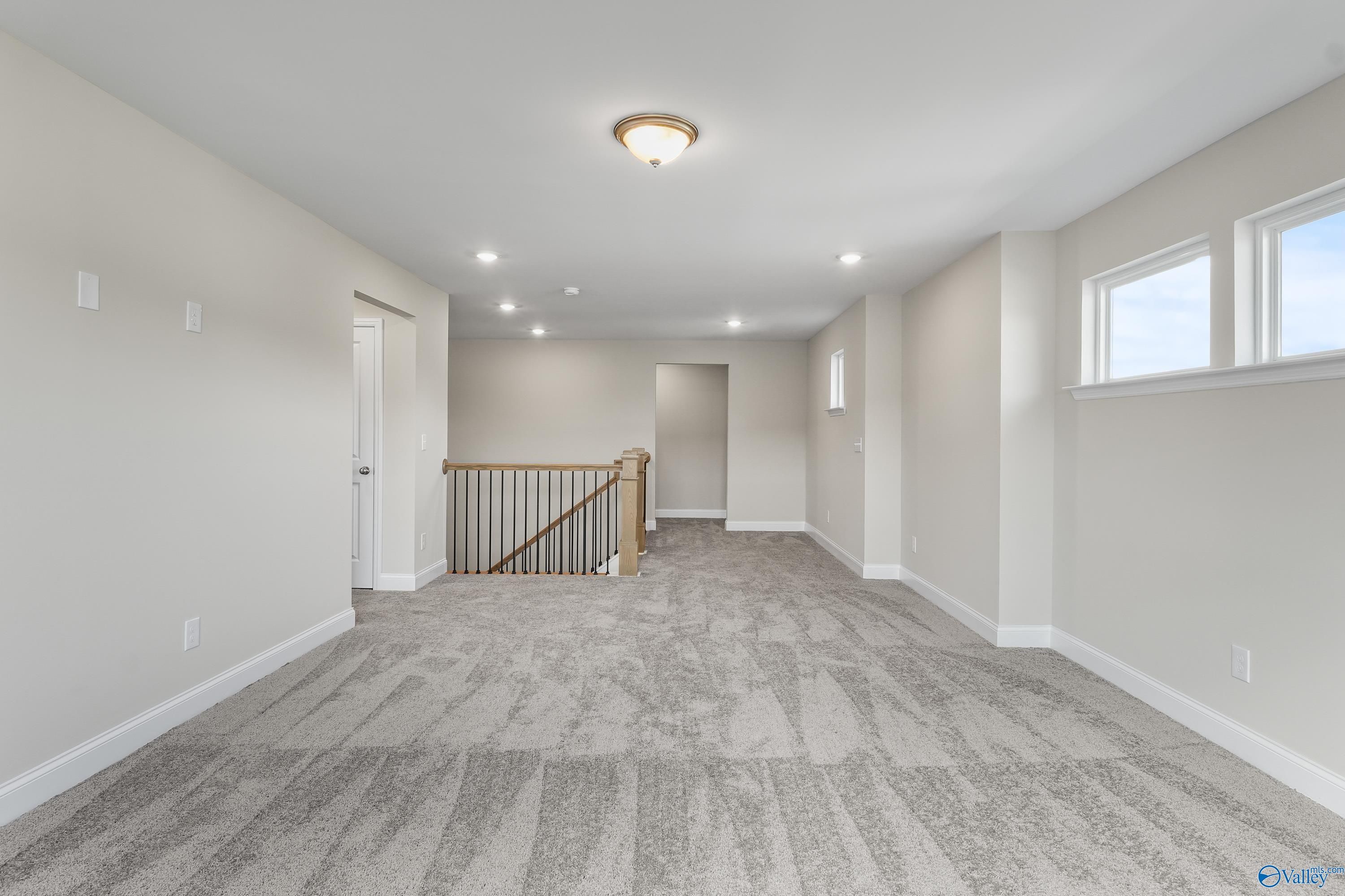 Spacious upstairs loft with beige carpet, wooden staircase railing, and large windows in Evermore Homes The Haven, Owens Cross Roads, Alabama