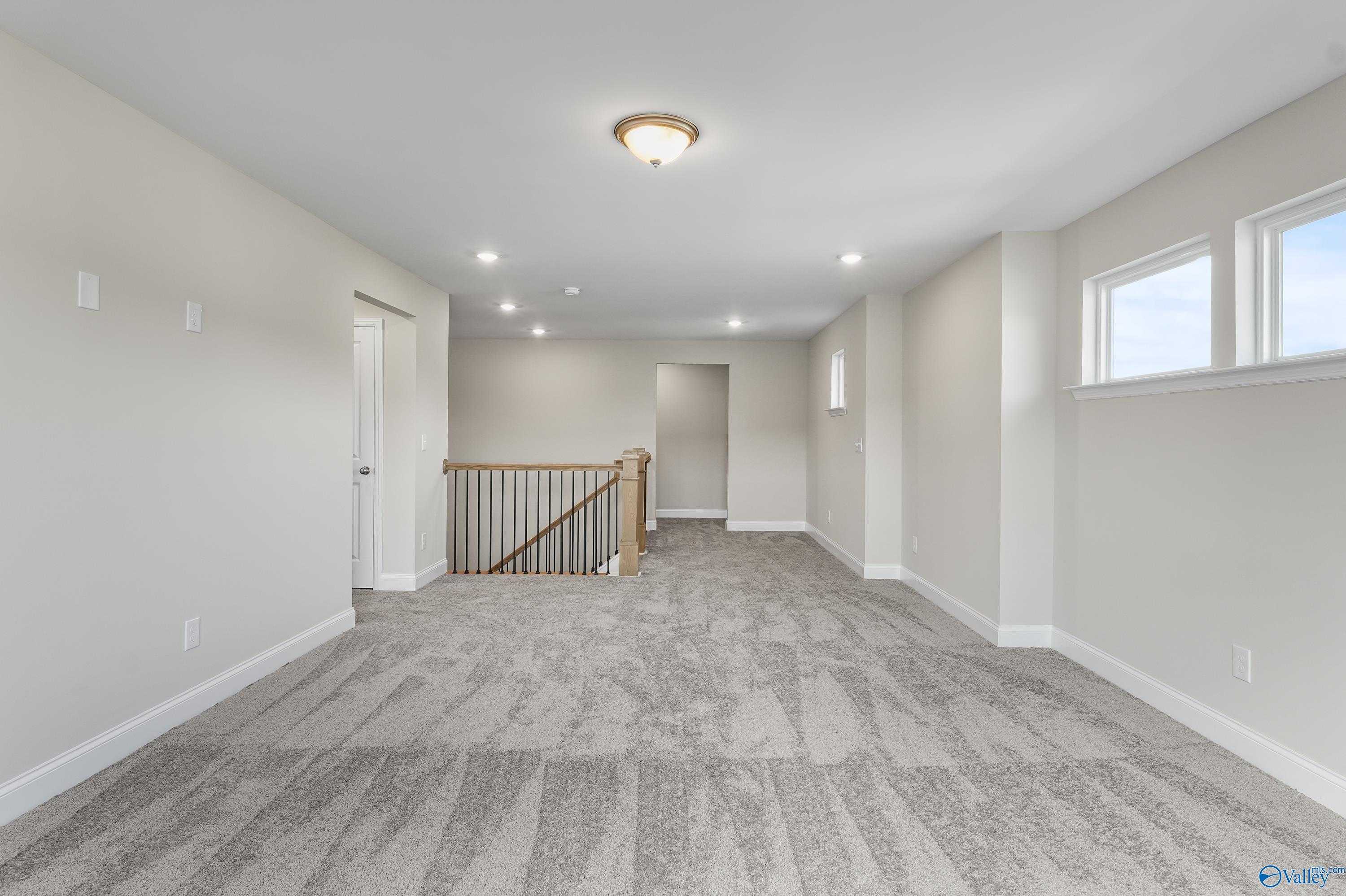 Spacious upstairs loft with beige carpet, wooden staircase railing, and large windows in Evermore Homes The Haven, Owens Cross Roads, Alabama