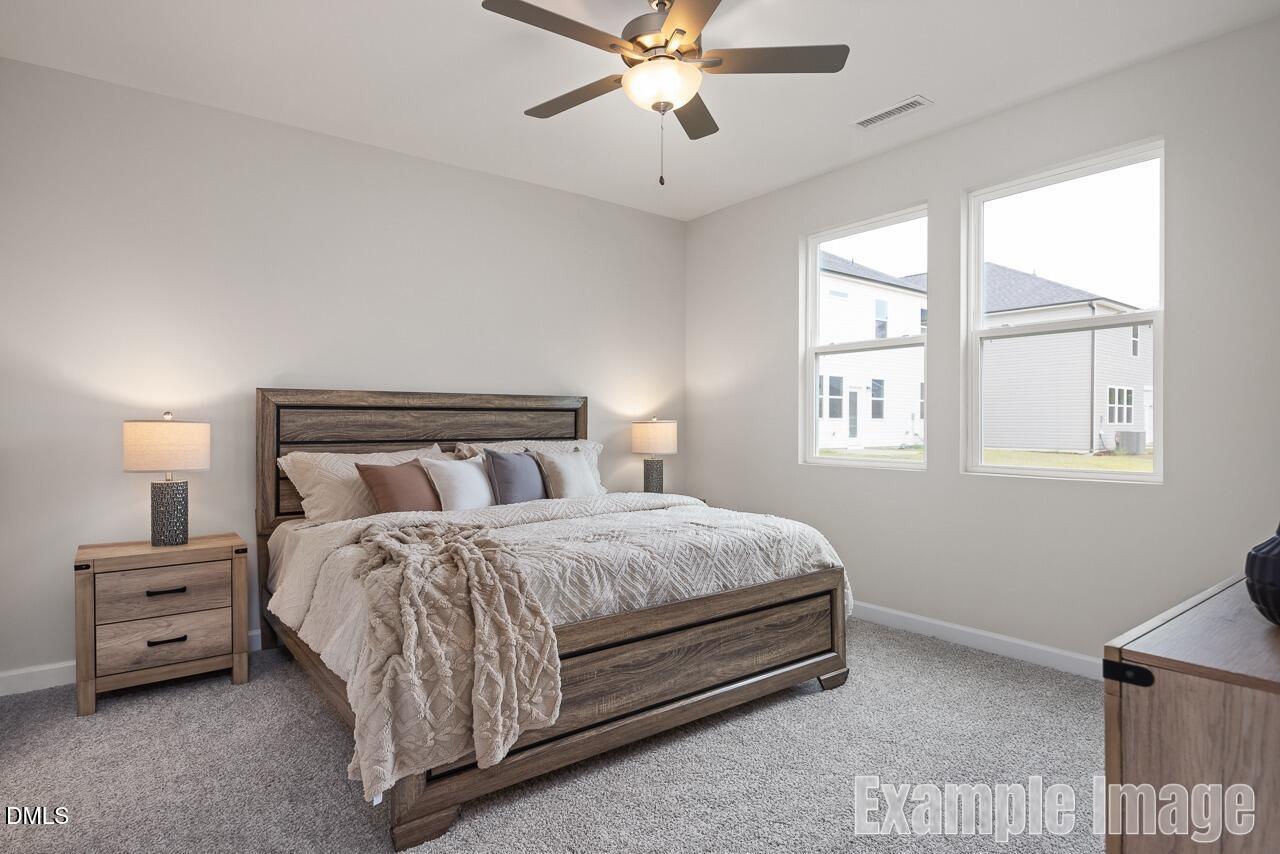 Cozy master bedroom with wooden bed frame, plush beige bedding, ceiling fan, and large windows in Davidson Homes The Carter C, Lillington, NC