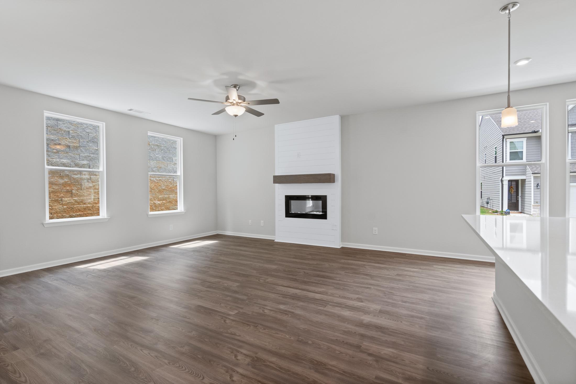 Open-concept great room in The Cary A with white fireplace, hardwood floors, large windows, and ceiling fan