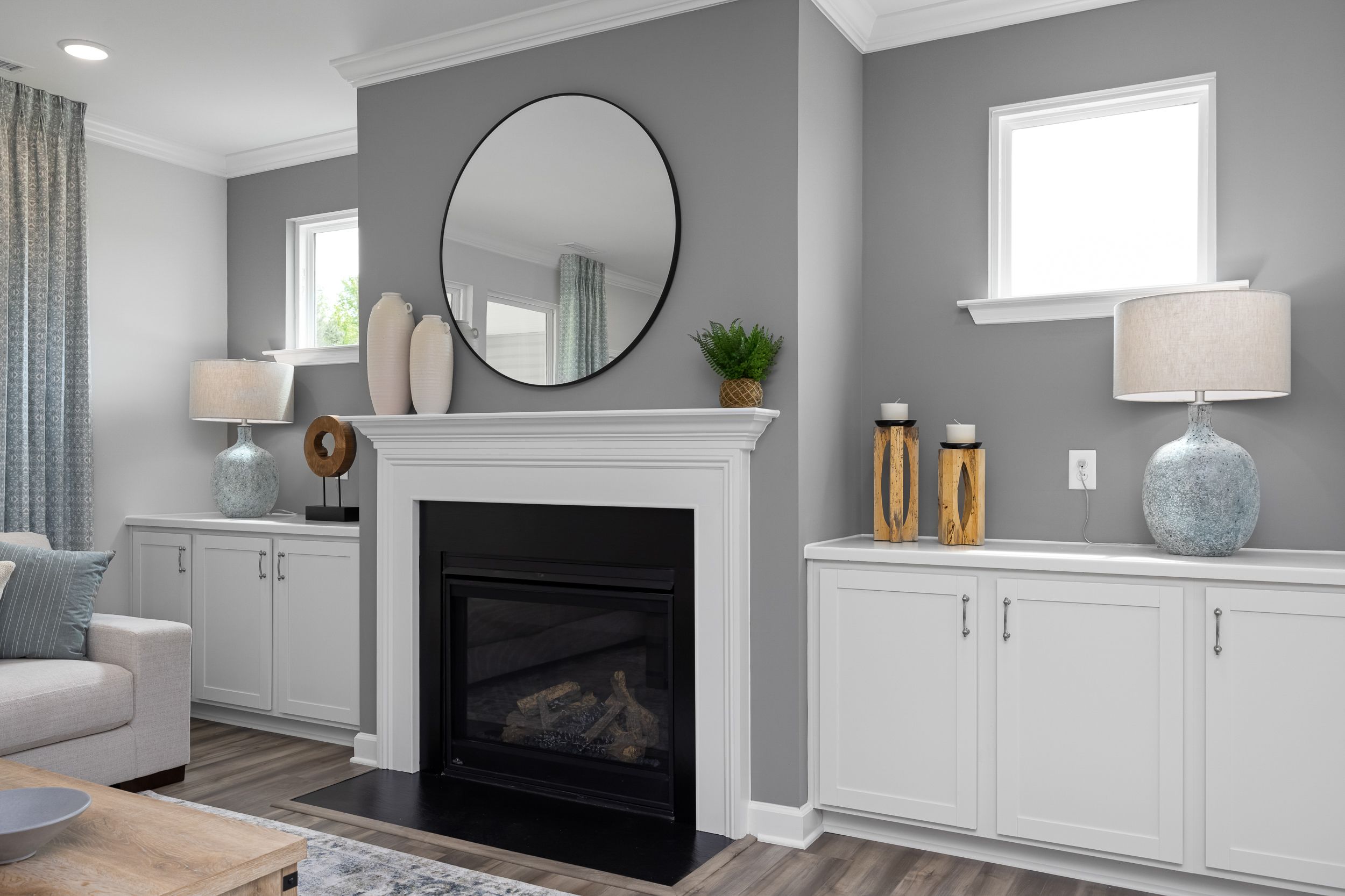 Cozy living room in The Magnolia B with gray walls, round black mirror, gas fireplace, and white built-in cabinets
