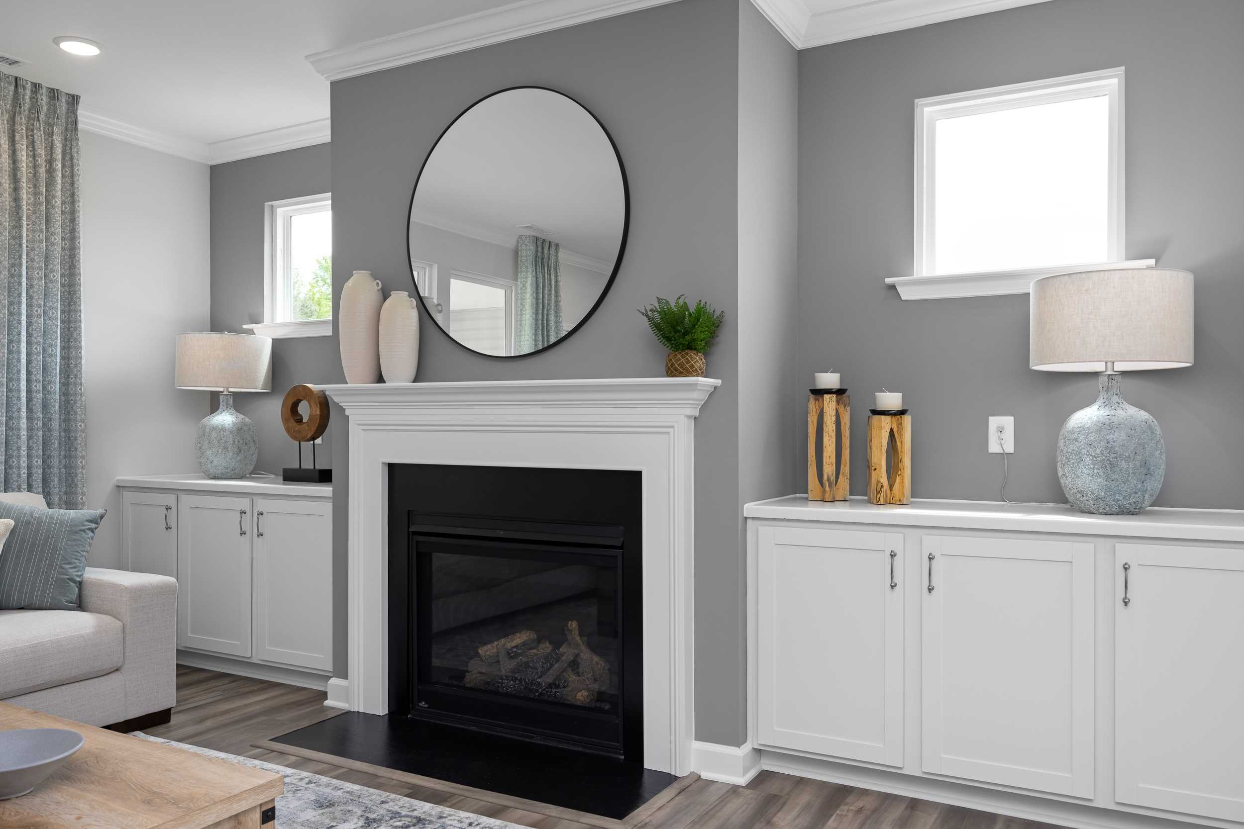 Cozy living room in The Magnolia A with gray walls, round black mirror, white built-in cabinets, and gas fireplace by Davidson Homes