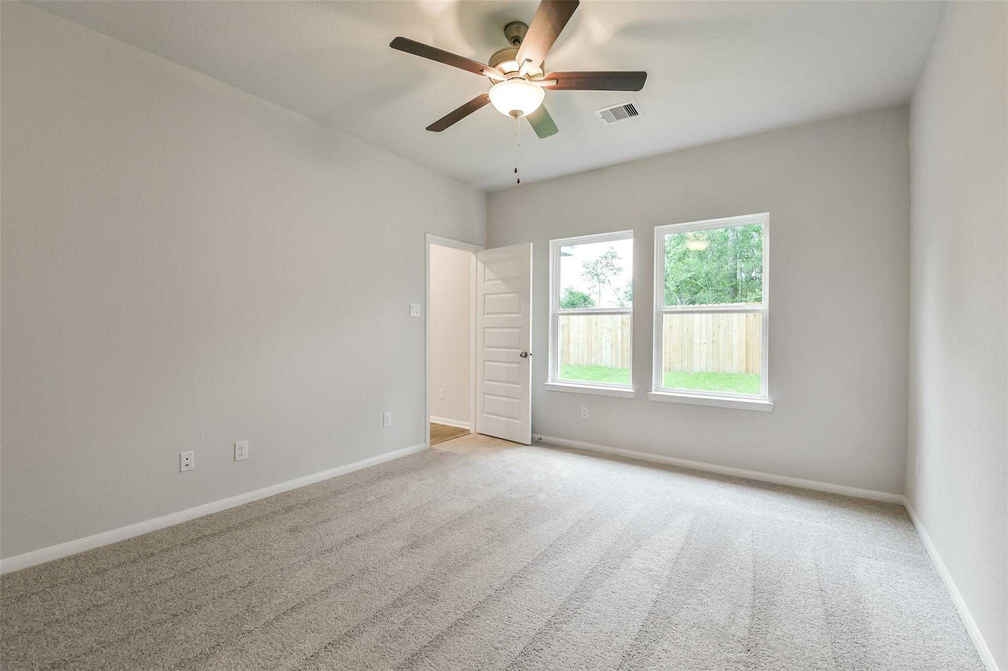Bright bedroom with ceiling fan, large windows overlooking fenced backyard, beige walls and carpet in Davidson Homes The Brazos F, Conroe Texas