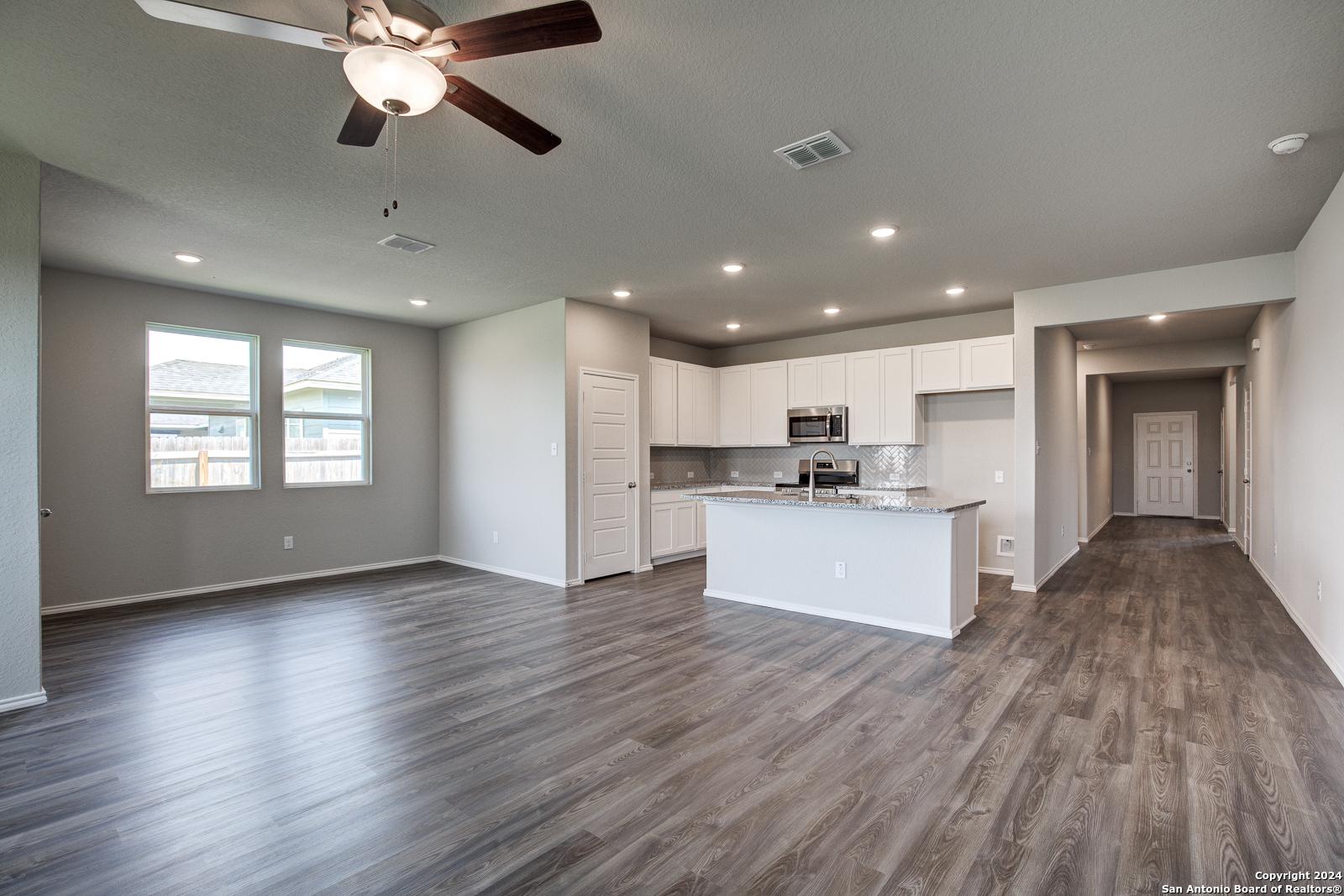 Open-concept kitchen and living area with white island cabinets, stainless appliances, ceiling fan, and wood floors in The Daphne J, Seguin, TX