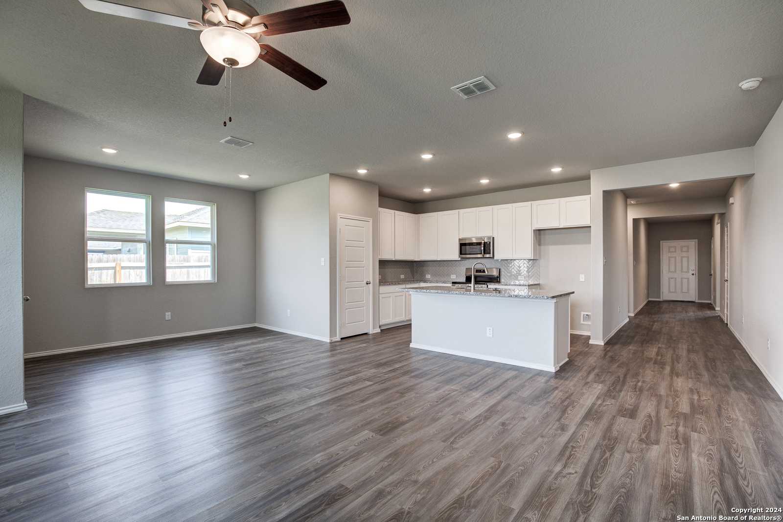 Open-concept kitchen and living area with white island cabinets, stainless appliances, ceiling fan, and wood floors in The Daphne J, Seguin, TX
