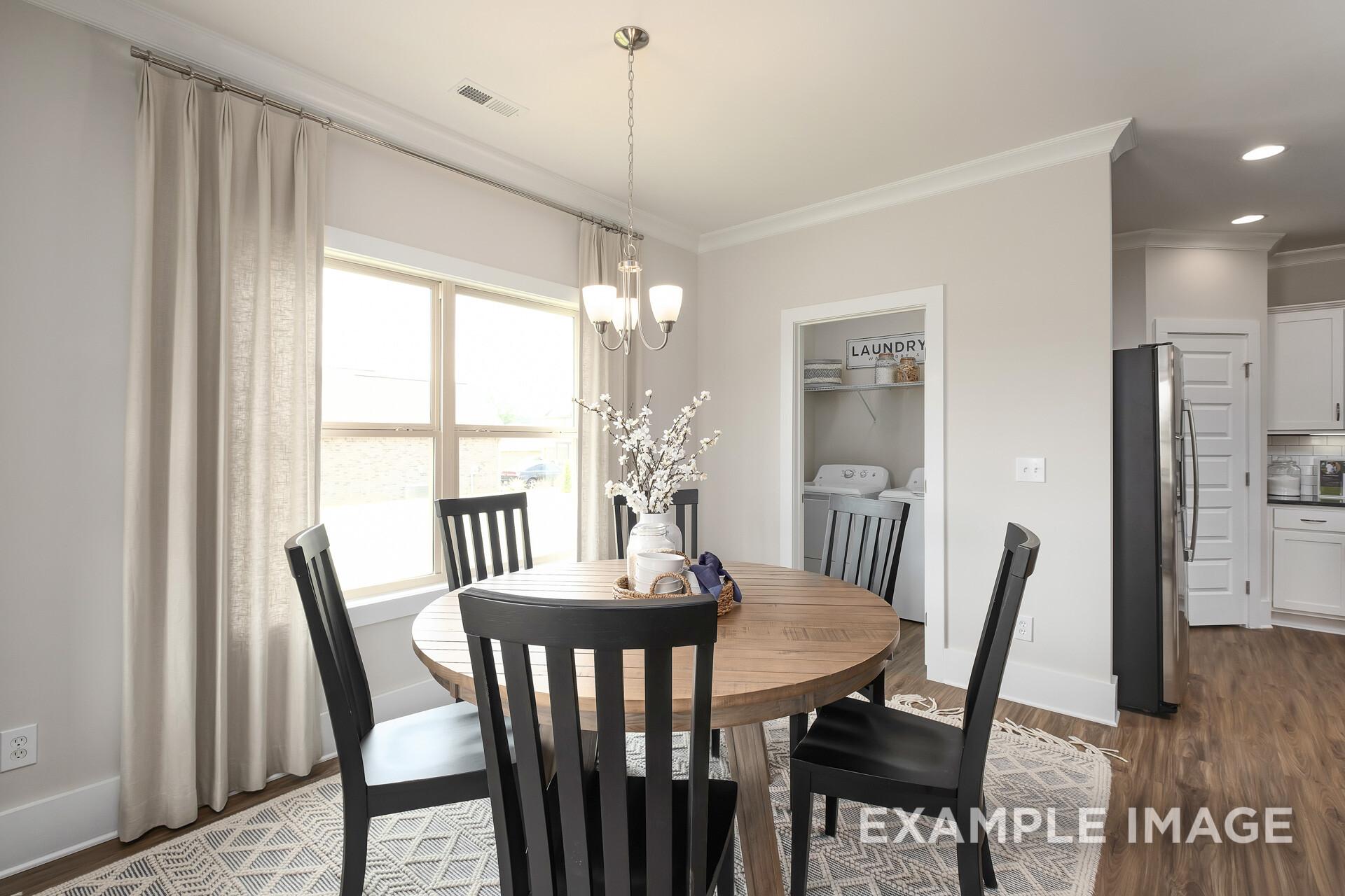 Dining room in The Daphne D home with round wooden table, black chairs, chandelier, sheer curtains, and adjacent laundry-kitchen area