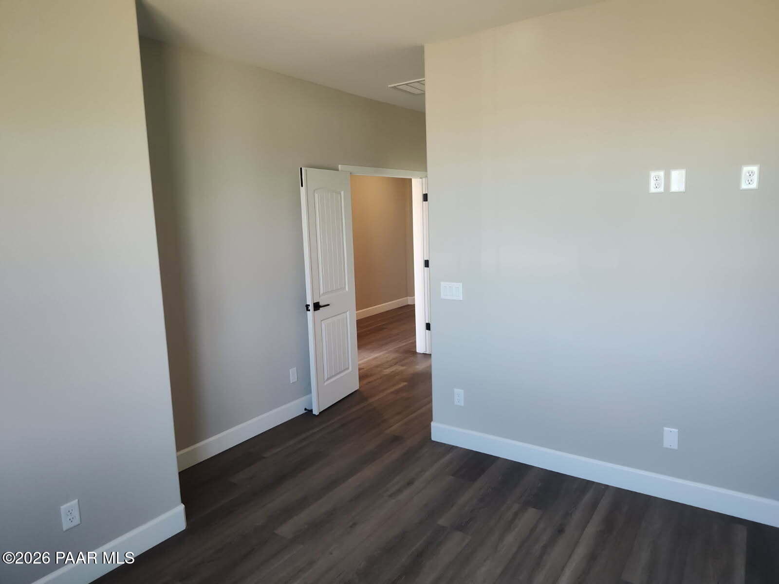 Spacious empty bedroom with beige walls, dark laminate flooring, open white door to hallway in Davidson Homes The Harmony C, Prescott Valley