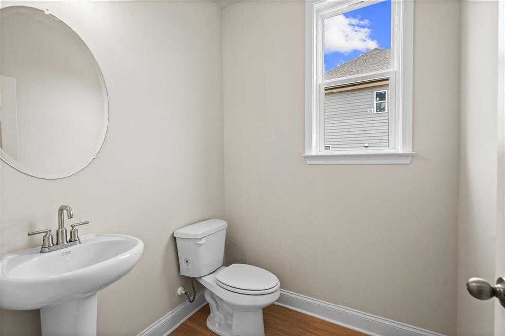 Bright powder room with round mirror, pedestal sink, toilet, and window view in Davidson Homes The Hickory B at Wehunt Meadows, Hoschton