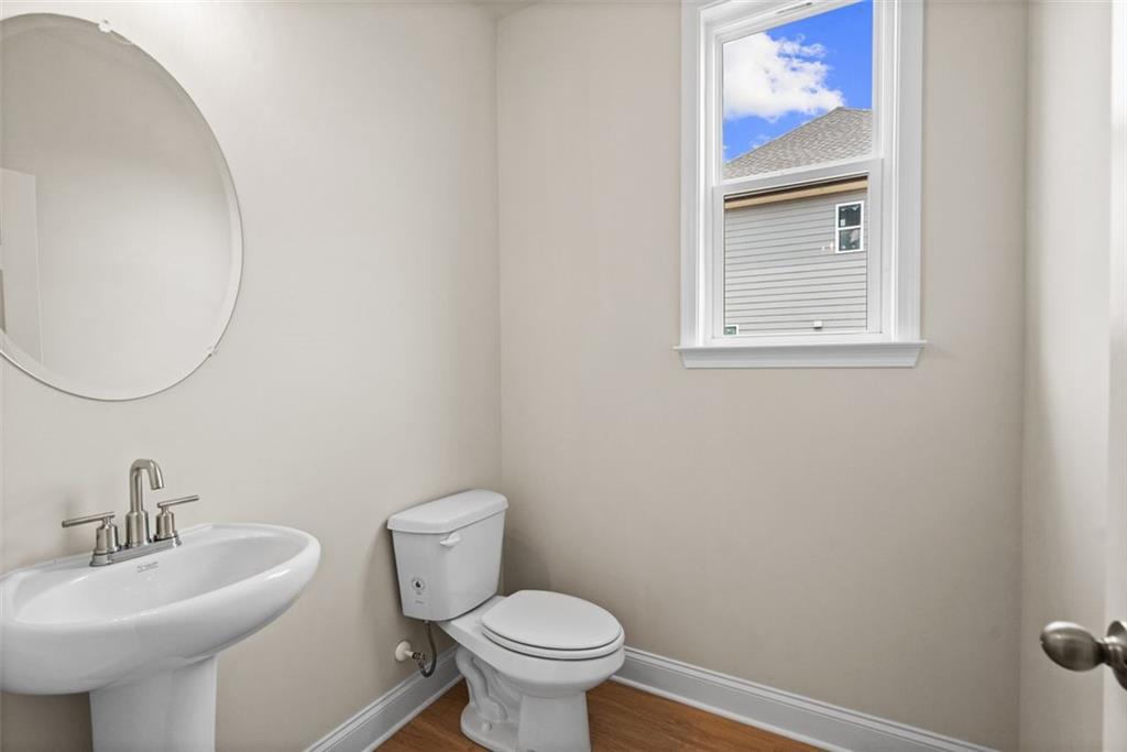 Bright powder room with round mirror, pedestal sink, toilet, and window view in Davidson Homes The Hickory B at Wehunt Meadows, Hoschton
