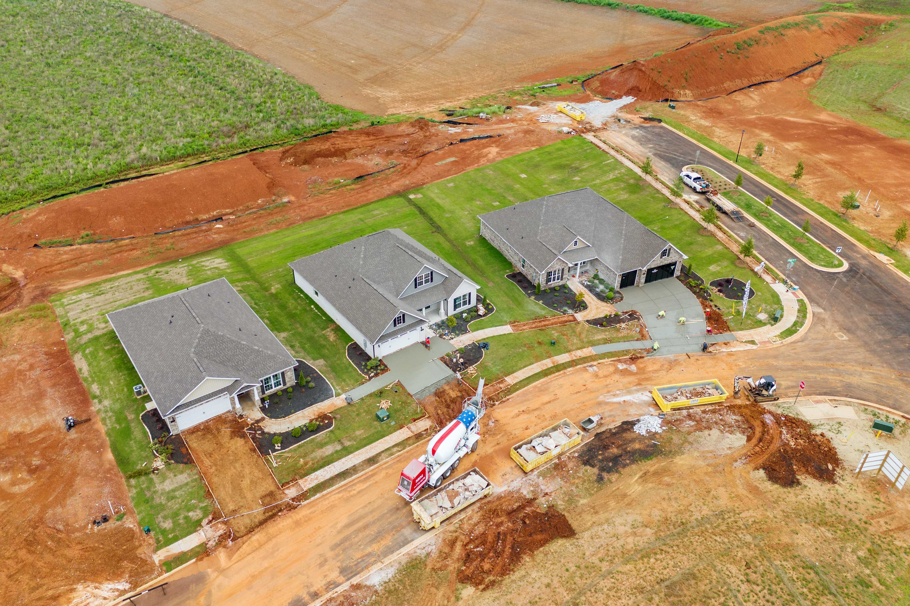 Aerial view of three new homes under construction at Creekside in Harvest, Alabama with concrete mixer truck and red clay fields