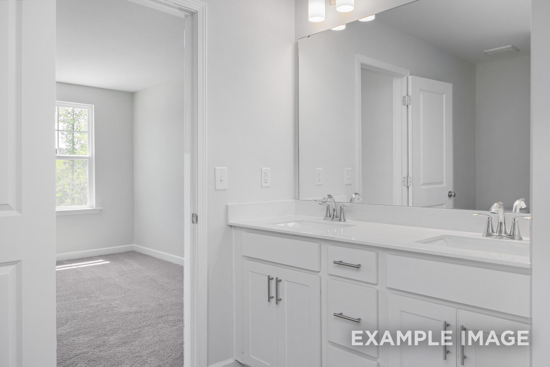 Spacious master bathroom in The Gavin B with white double vanity, large mirror, and open view to carpeted bedroom