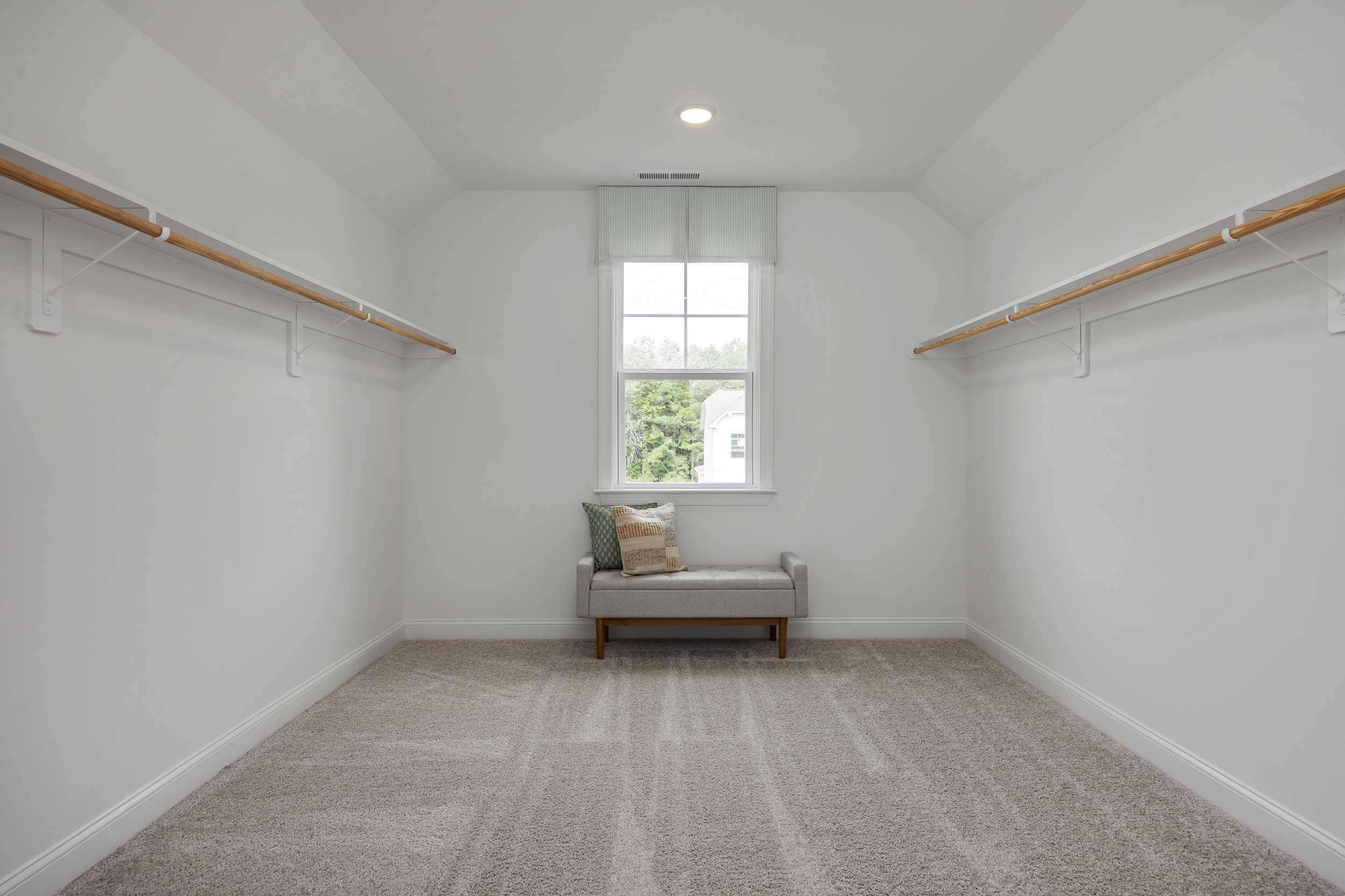 Spacious walk-in closet at Laneridge Estates in Raleigh NC featuring white walls, wooden hanging rods, window, bench, and carpeted floor