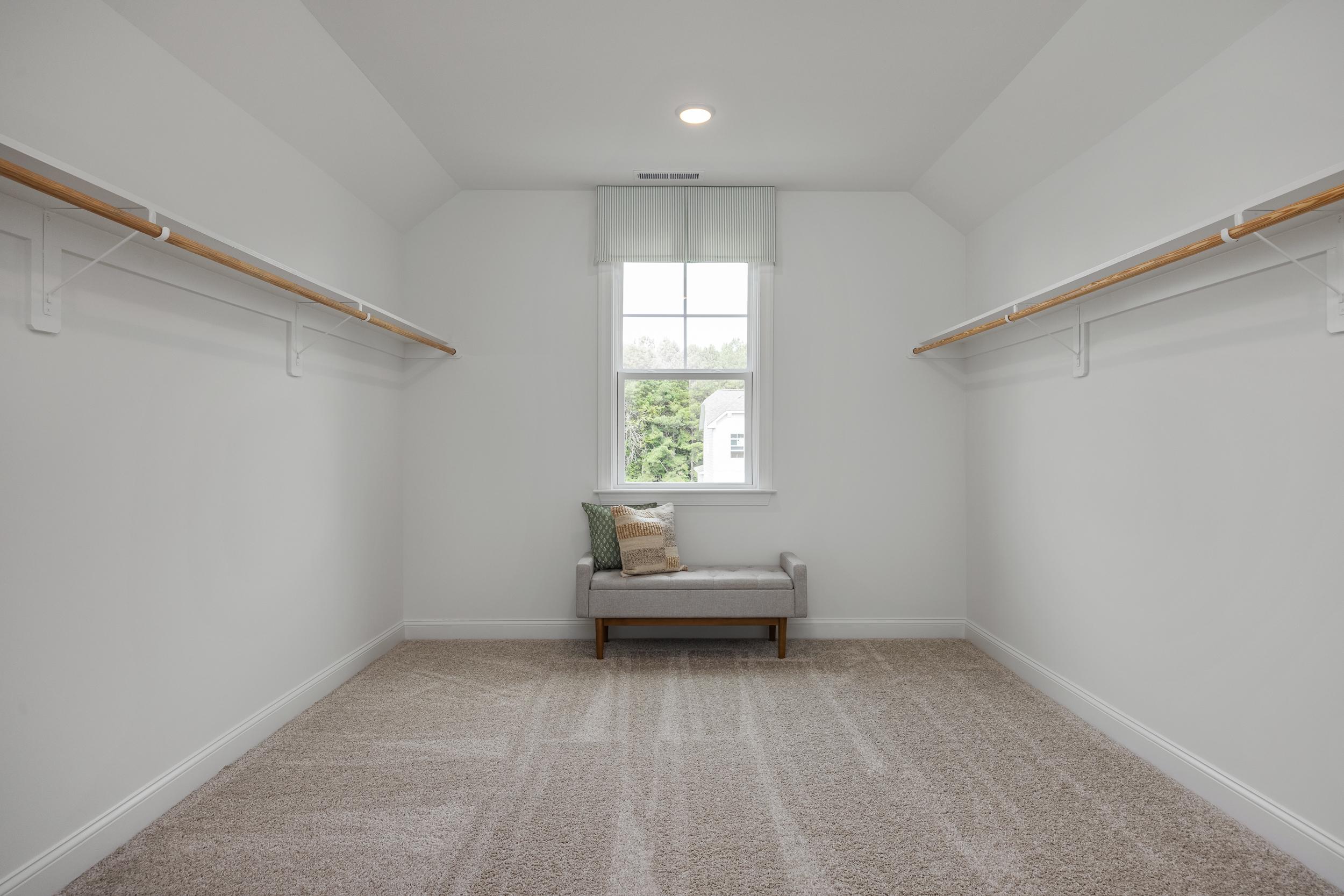 Spacious walk-in closet at Laneridge Estates in Raleigh NC featuring white walls, wooden hanging rods, window, bench, and carpeted floor