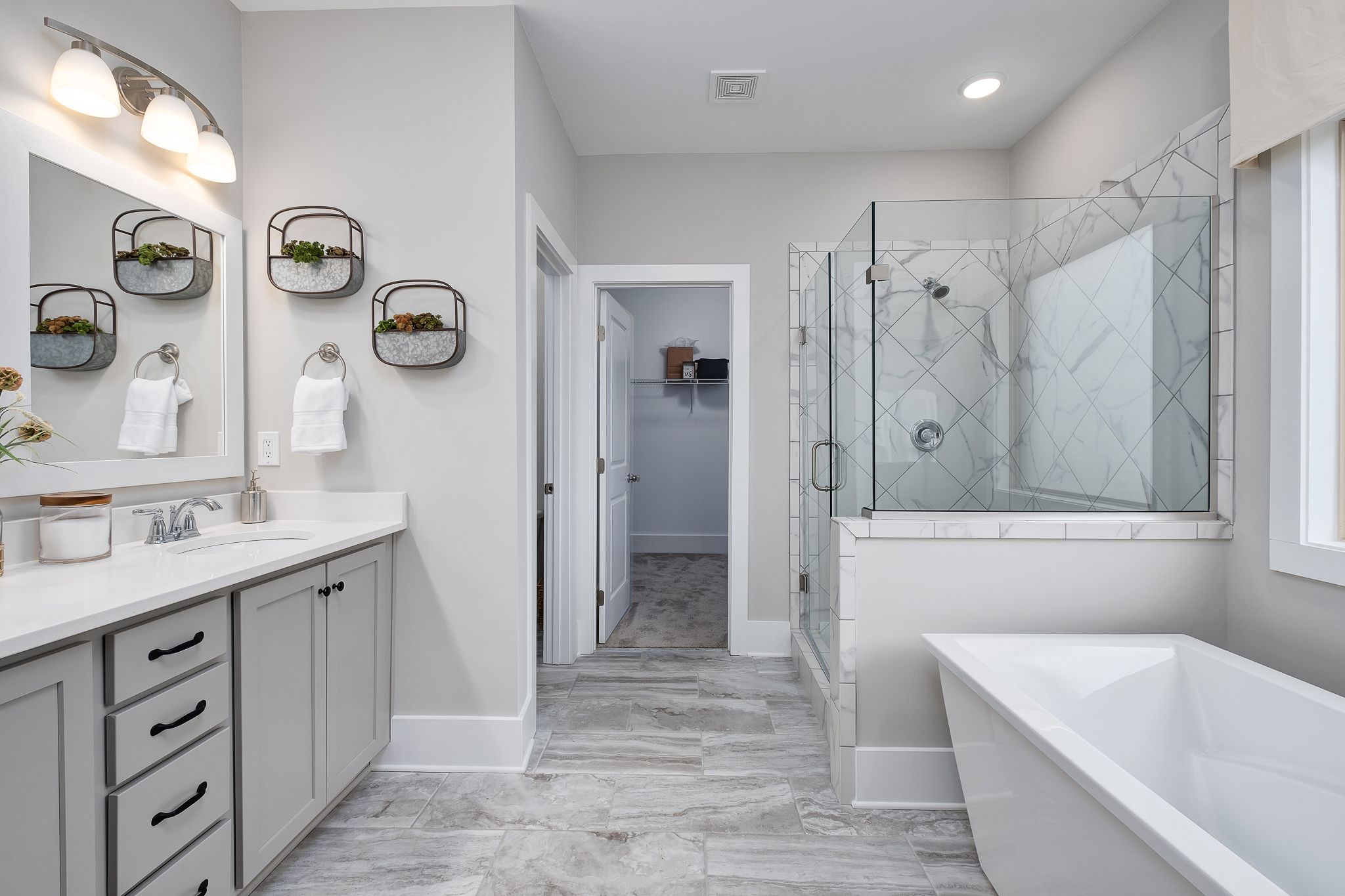 Spacious master bathroom in Chimney Creek Hampton Cove AL with freestanding tub, glass shower, double vanity, and gray cabinetry by Davidson Homes