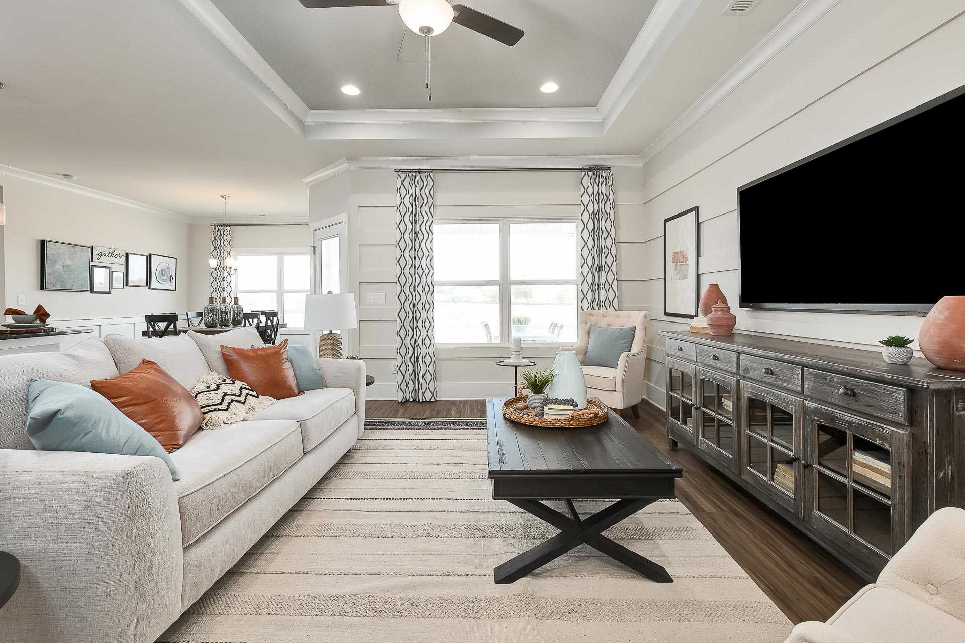 Open-concept living room in Newby Chapel Athens Alabama with beige L-shaped sofa, rustic wood coffee table, wall-mounted TV, and sheer window curtains